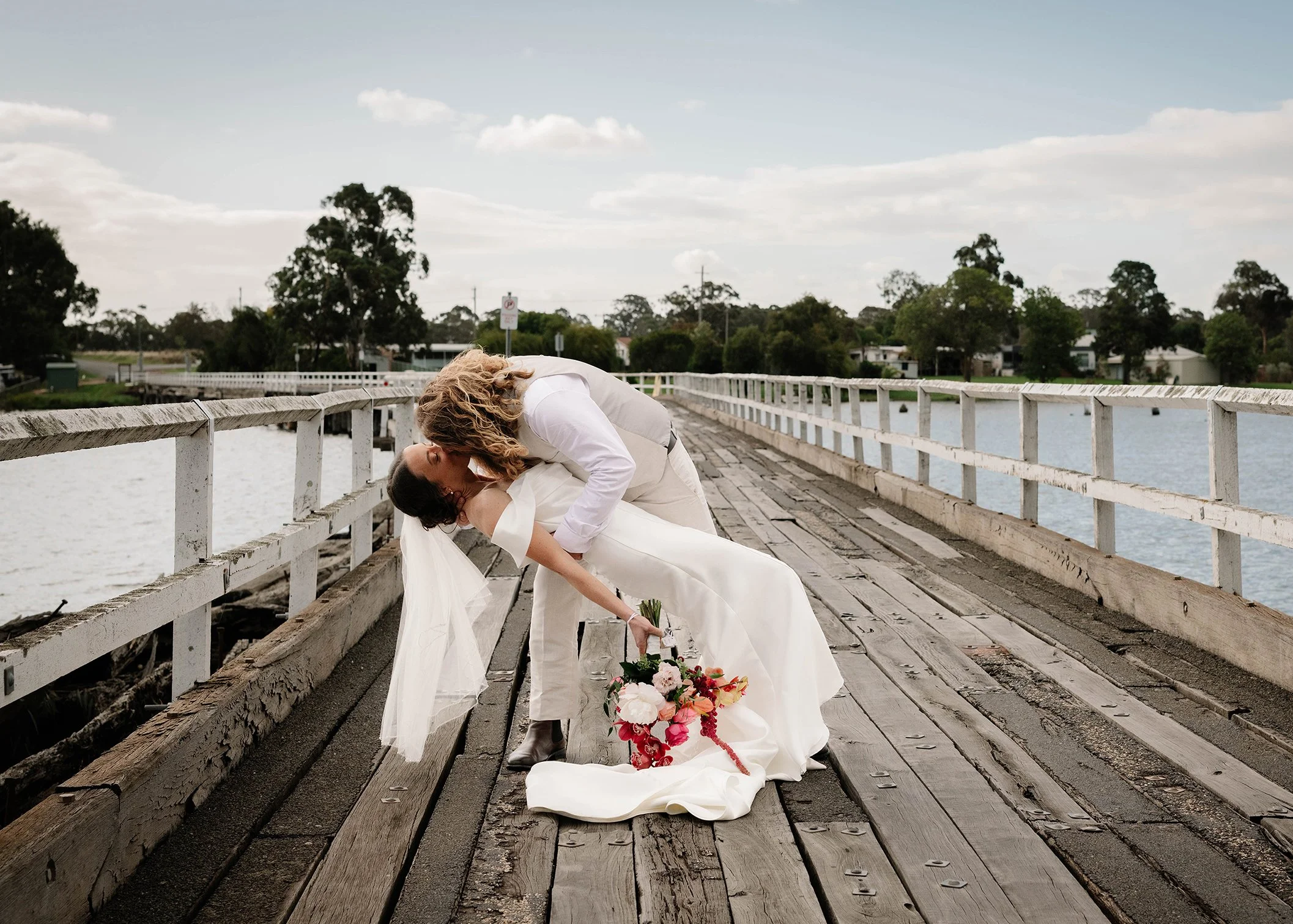 A couple dressed in wedding attire sharing a kiss on a wooden pier by the water, with a bouquet of flowers on the ground nearby.