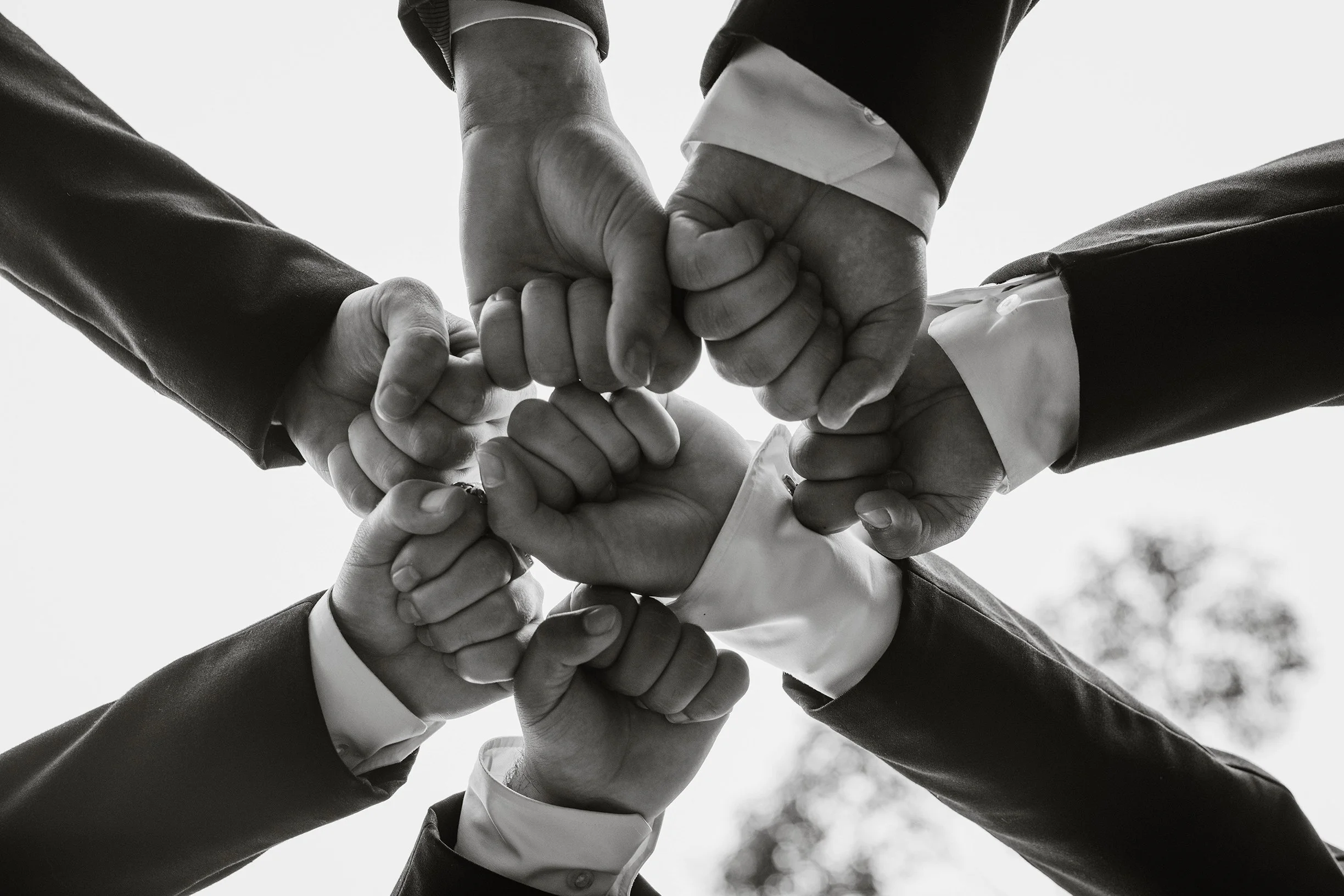 Seven people in business suits putting their fists together in a circle, symbolizing teamwork and unity, in black and white.