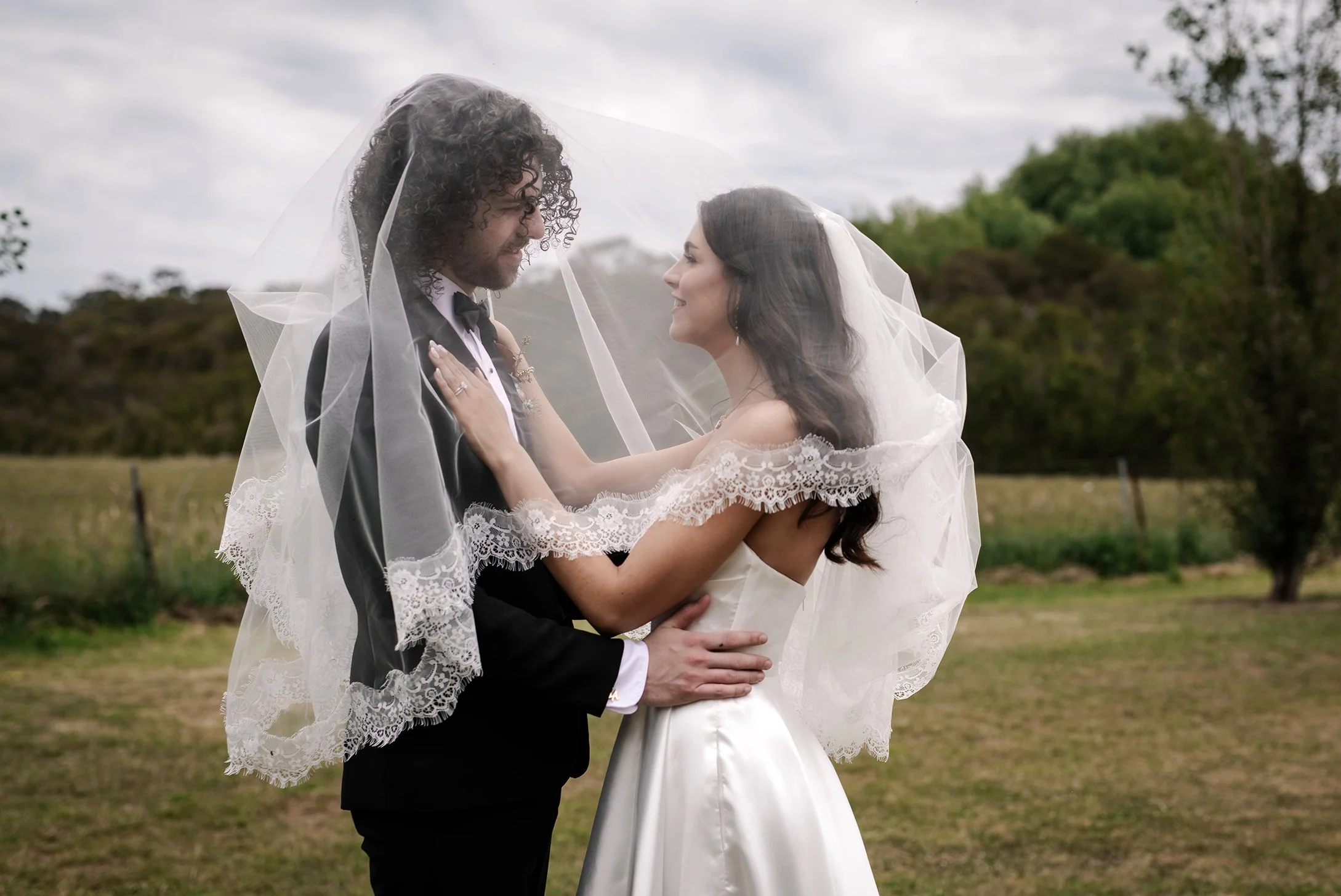 A bride and groom face each other outside, under a veil, with nature in the background.
