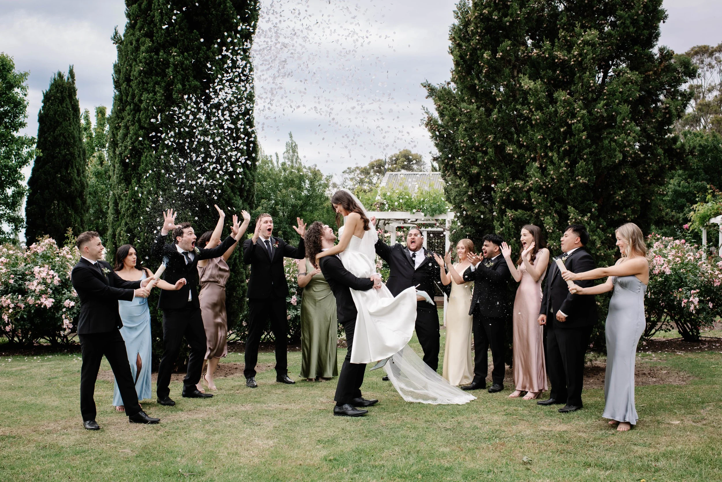 A wedding celebration outdoors with a bride and groom lifting each other in front of a group of people throwing confetti and cheering, surrounded by tall trees and blooming bushes.