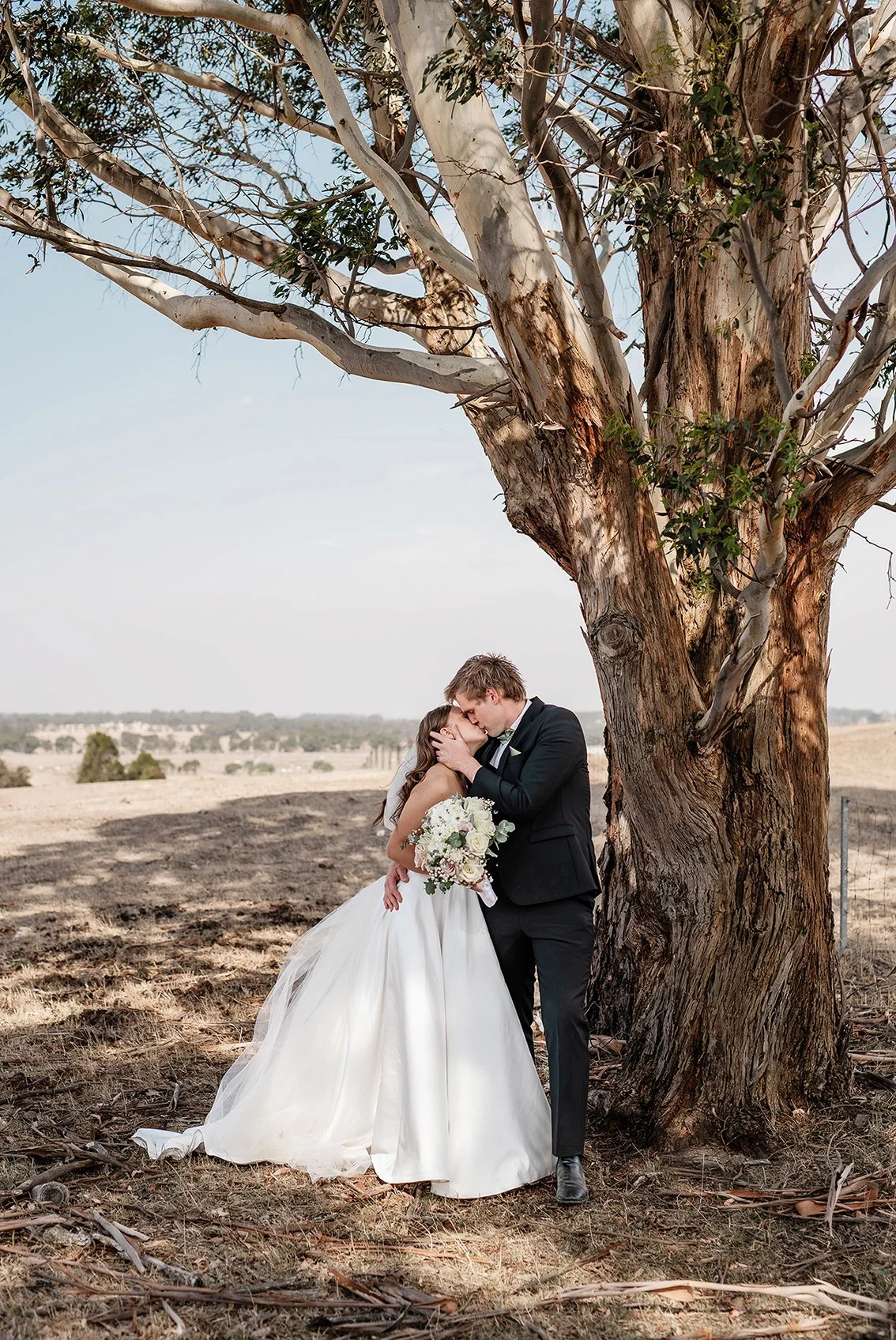 Bride holding a bouquet while the groom embraces her beneath a large gum tree during an outdoor wedding portrait