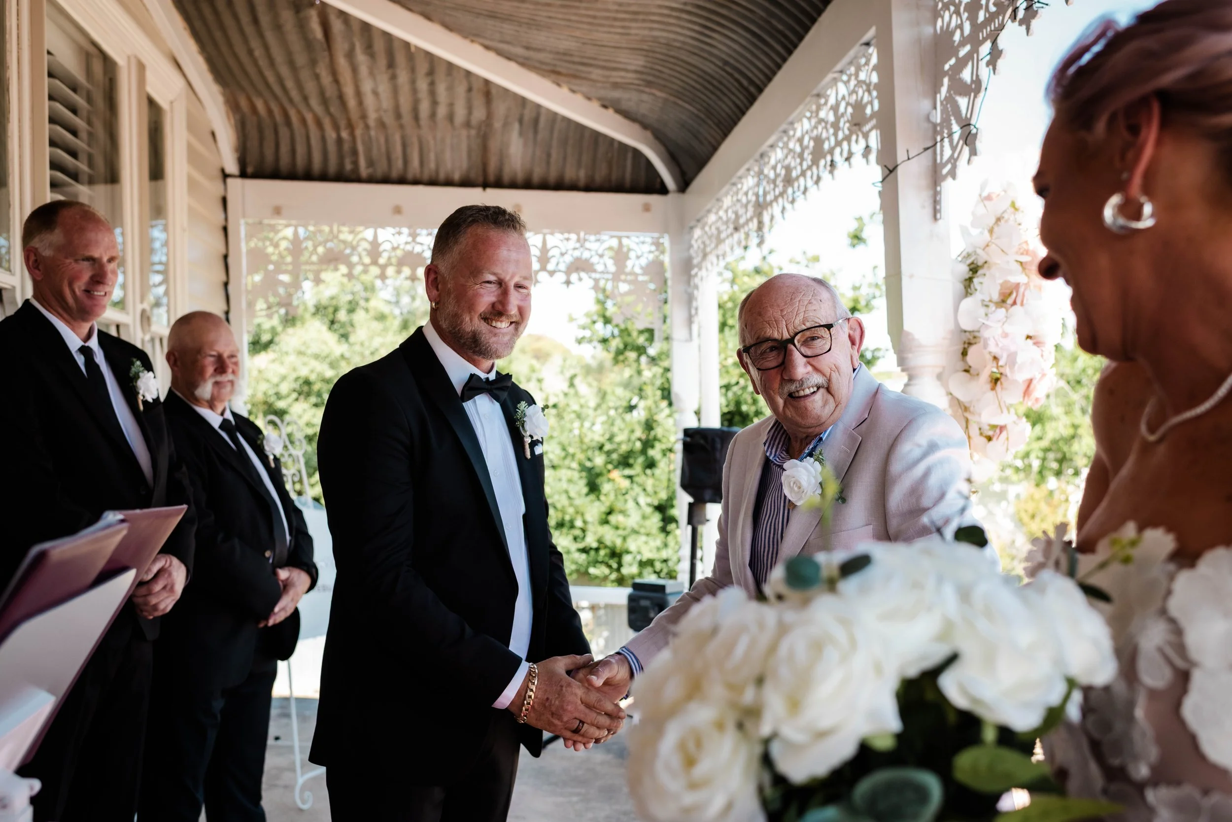 Groom in black tuxedo shaking hands with an elderly family member during an outdoor wedding ceremony with the bride nearby
