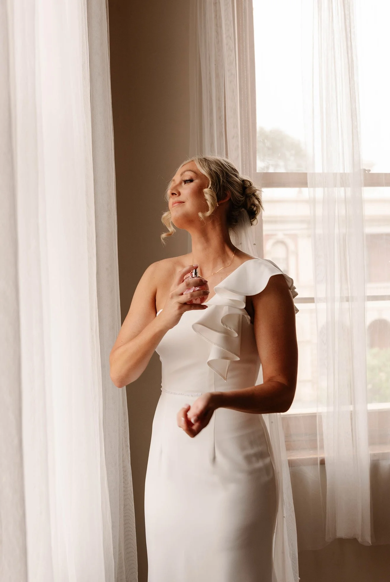 A woman in a white dress standing by a window, holding a perfume bottle, with sheer curtains and a cityscape visible outside.