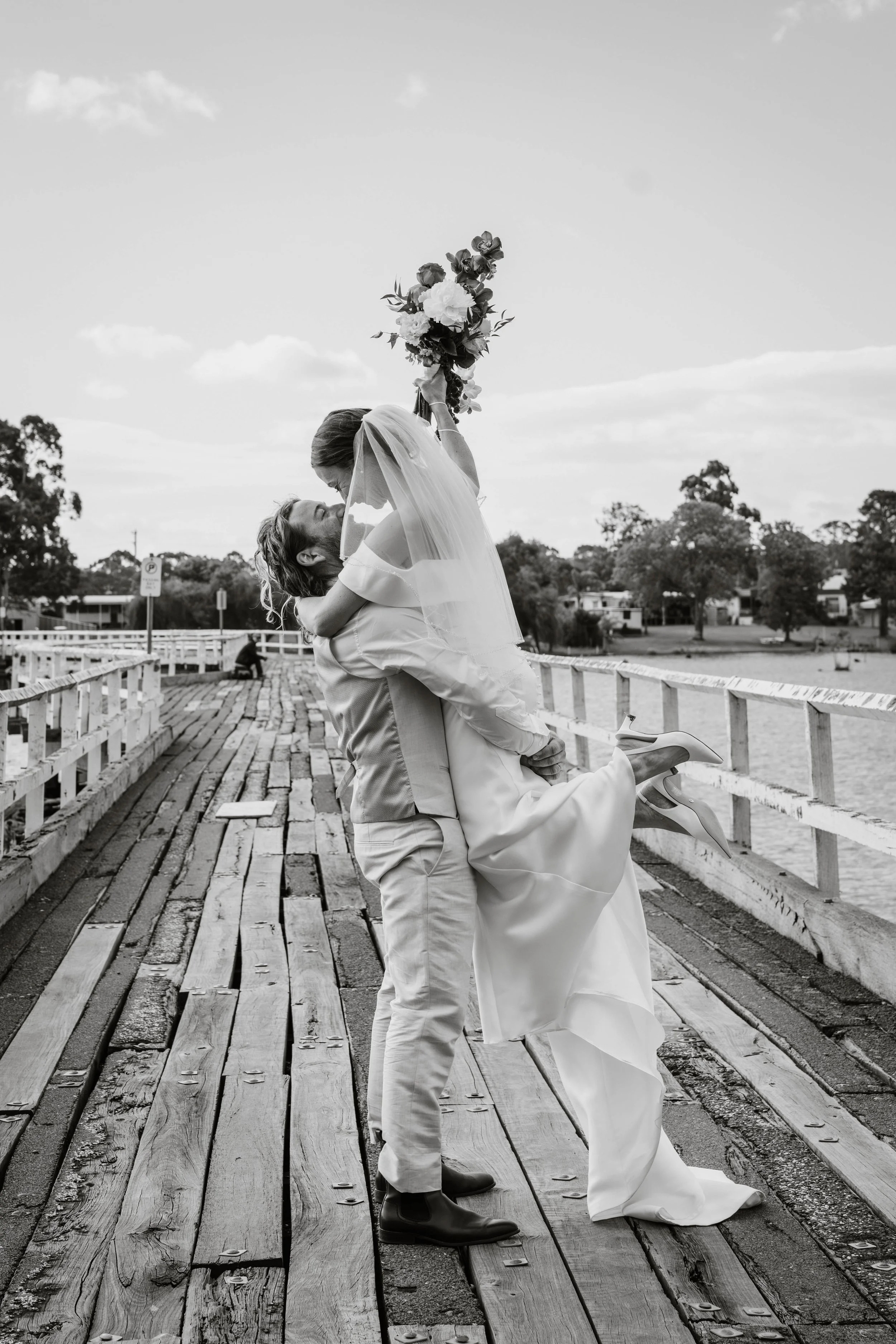 A couple, the man kneeling and lifting the woman off the ground, on a wooden pier with trees and houses in the background. The woman is holding a bouquet of flowers and is dressed in a wedding gown and veil, while the man is in a suit. The woman appears to be leaning in for a kiss.