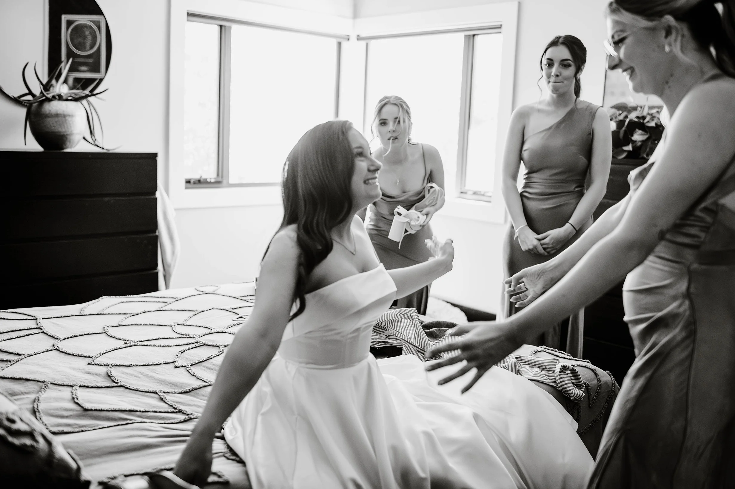 Candid photo of bride sitting on bed as bridesmaids all smile and reach to hug her