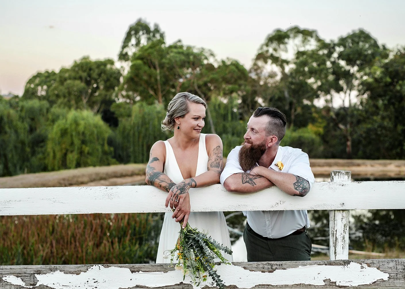 A couple with tattoos leaning on a white wooden fence, smiling at each other outdoors with green trees in the background.