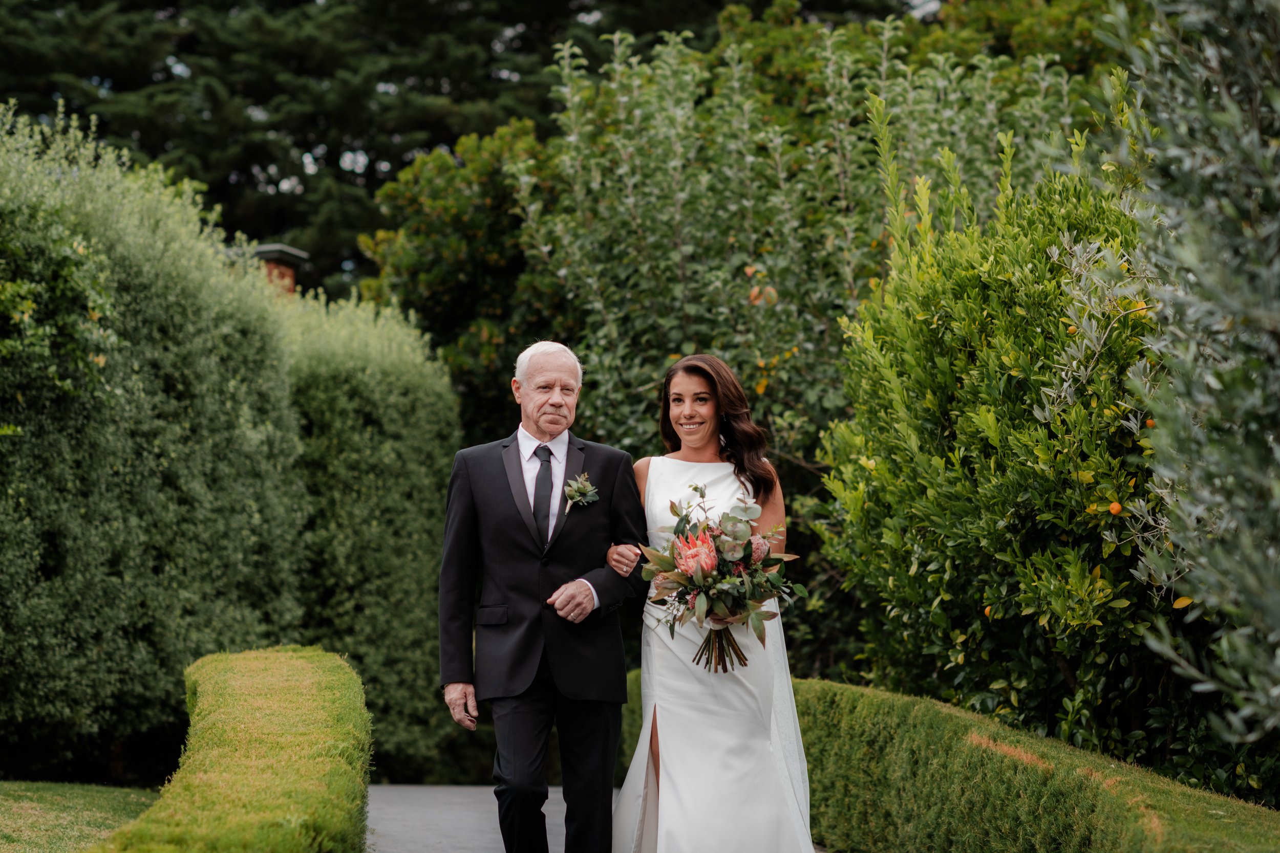 Father walking bride down a garden path lined with hedges at Jason's Restaurant Beaconsfield wedding ceremony