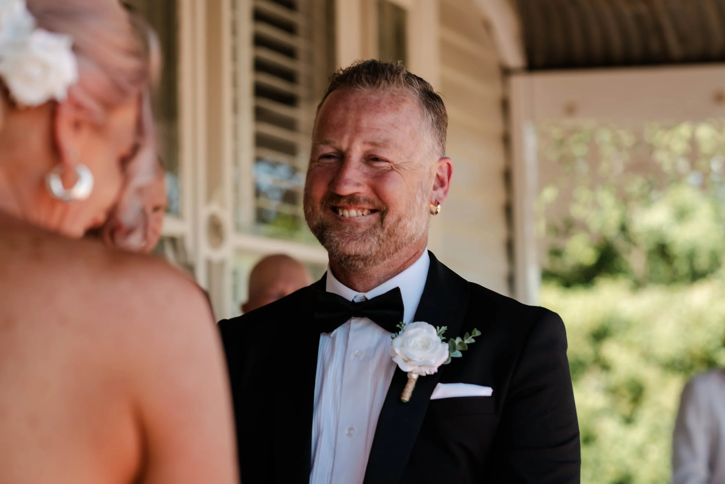 Groom in black tuxedo smiling at bride during an outdoor wedding ceremony on a covered veranda