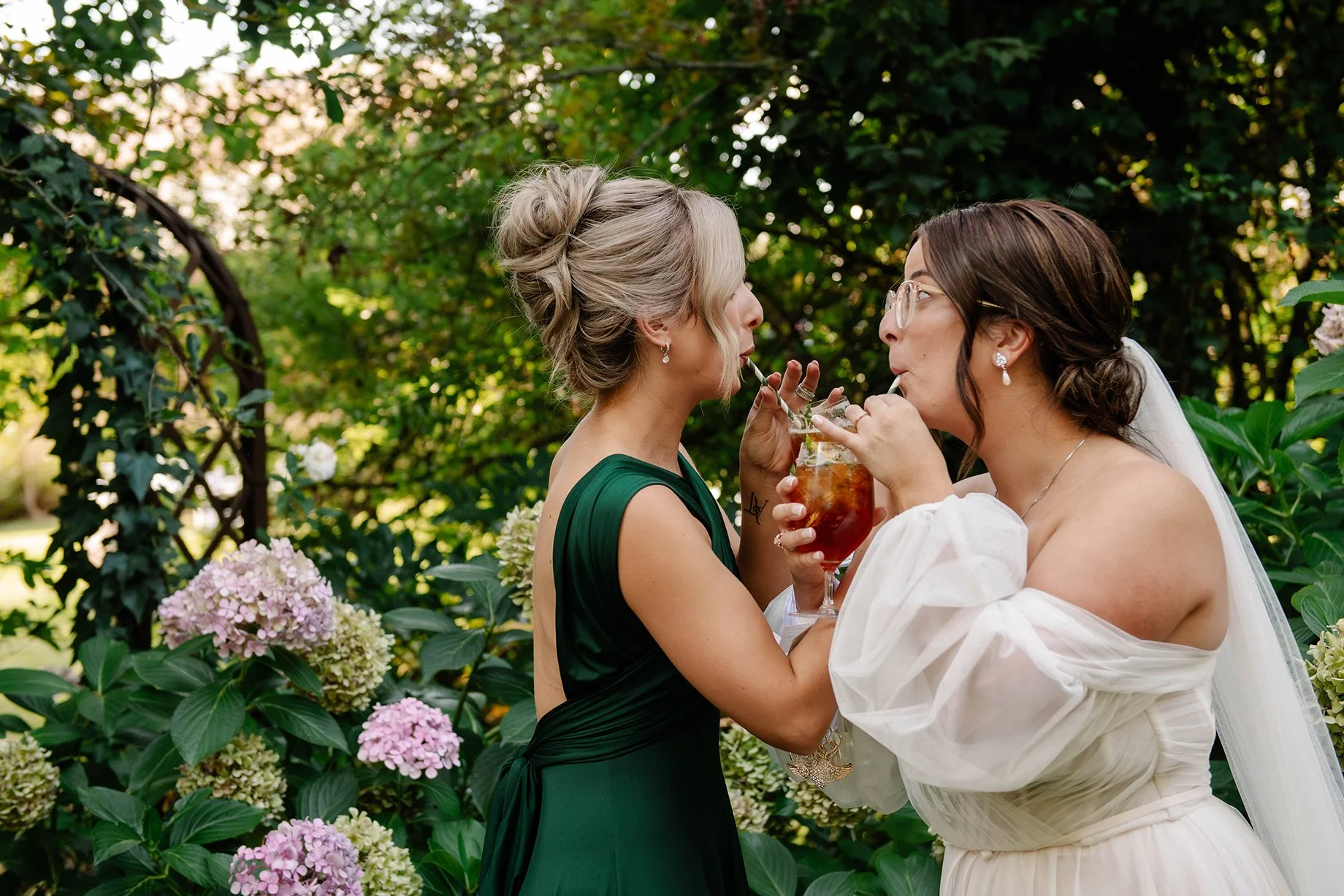 bride and maid of honor link arms and drink through straws from orange drinks in green garden in Bendigo