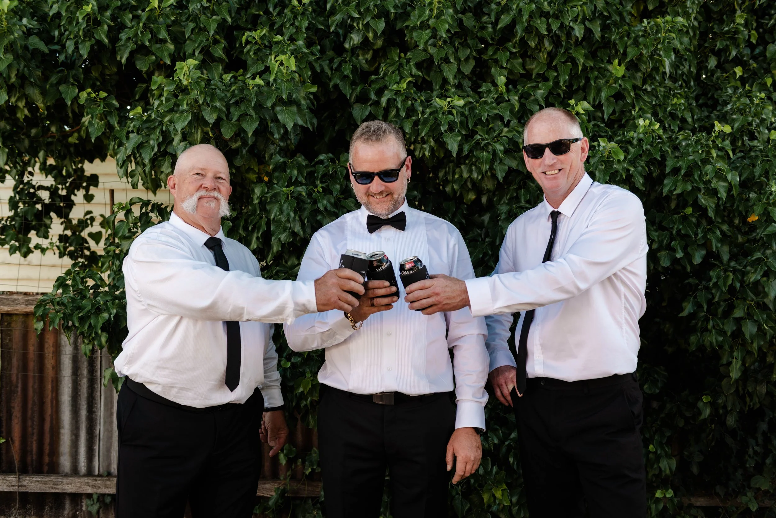 Three men in white shirts, black ties, and sunglasses raising drinks in front of a leafy green wall.