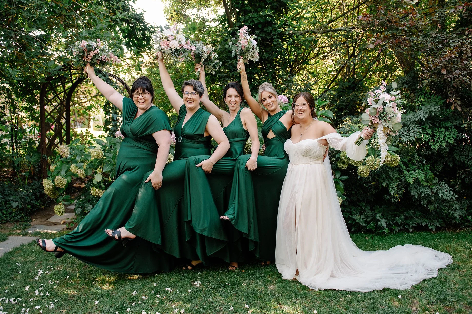 bridal party in green dresses with bride all hold their bouquets in the air and do funny pose holding each others raised leg in Bendigo