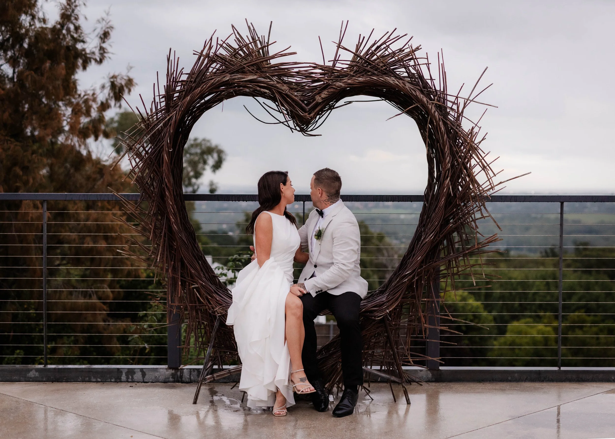 Bride and groom sitting together inside a heart-shaped twig arch on the terrace at Jason's Restaurant Beaconsfield with sweeping views behind them