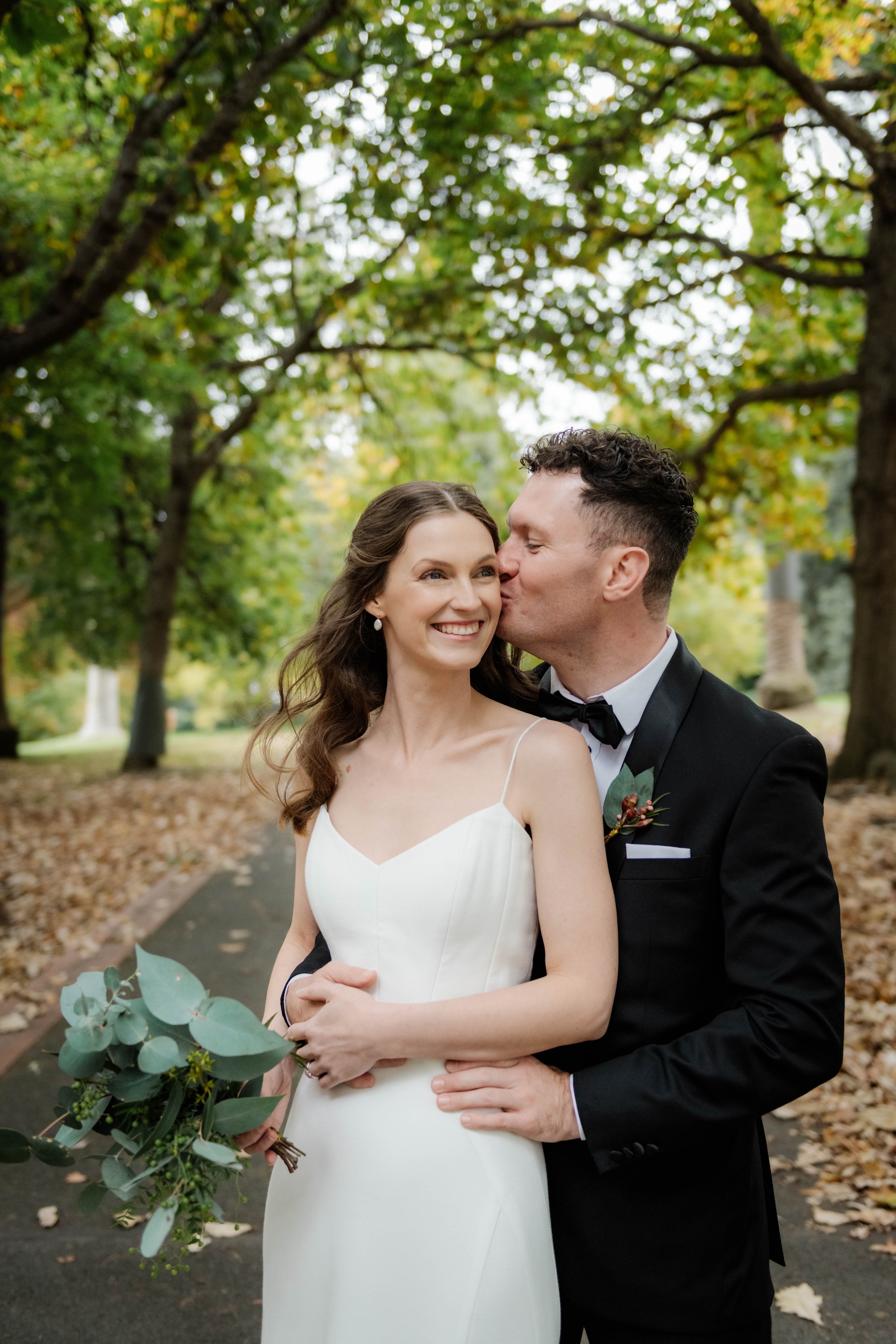 Bride and groom sharing a tender moment among autumn leaves in Fitzroy Gardens, Melbourne — candid wedding portrait by Kandi Lane Photography
