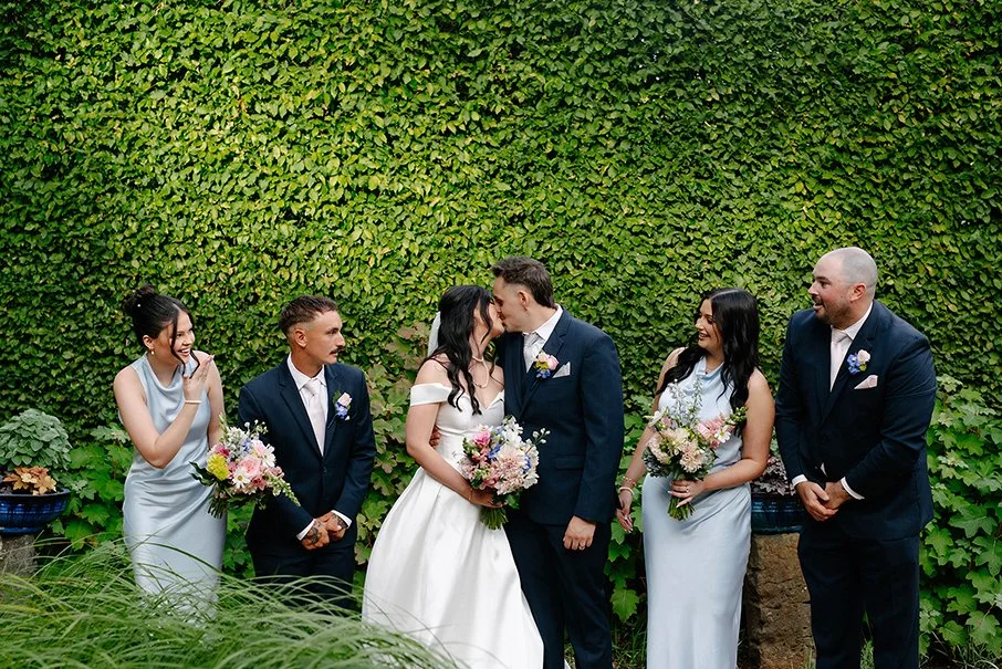 Wedding party kissing and laughing in front of a lush green hedge wall at Cloudehill Gardens Dandenong Ranges
