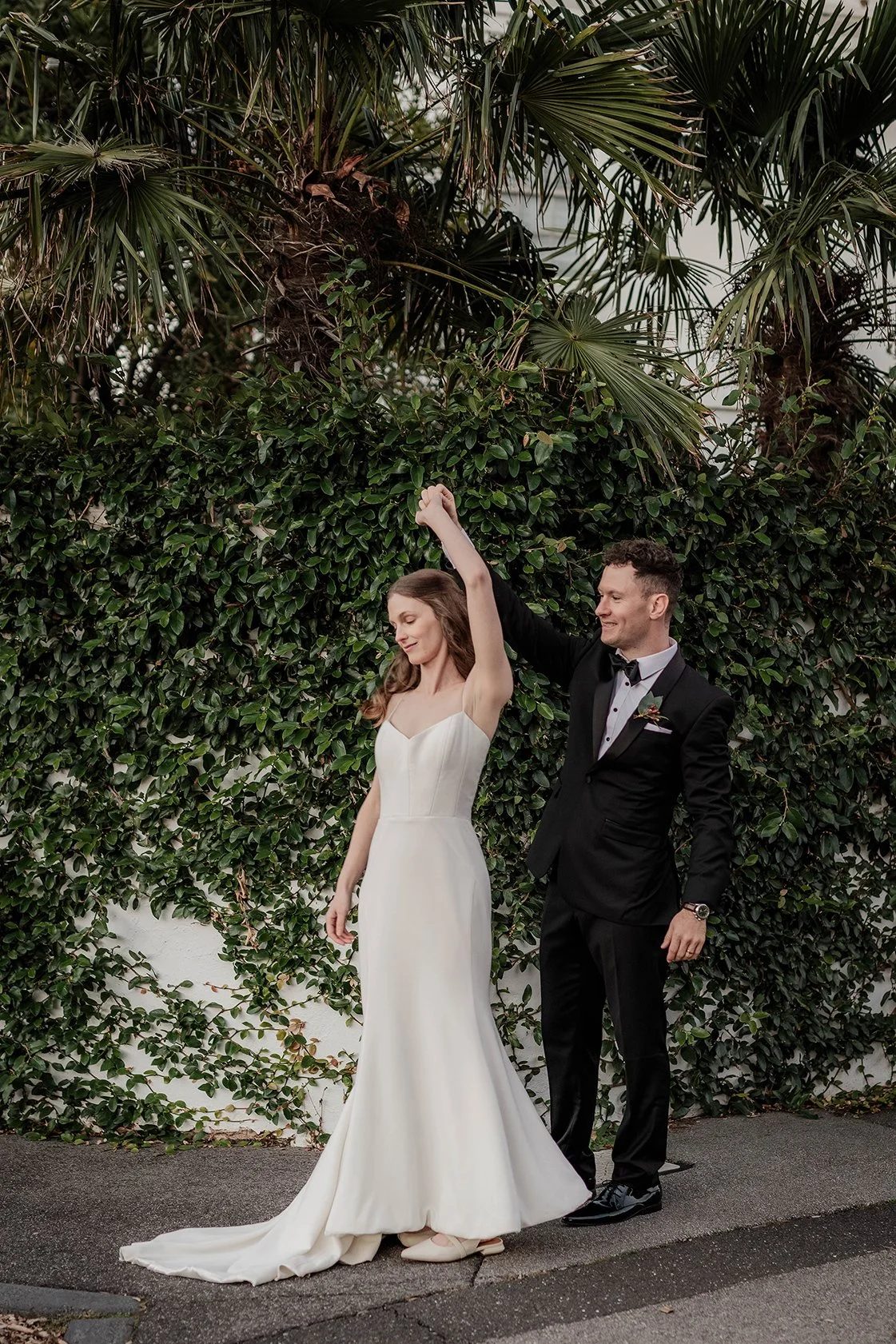 A bride and groom dancing outdoors against a green leafy hedge and palm trees. The bride wears a white strapless wedding gown and the groom wears a black tuxedo with a bow tie. They are holding hands, with the groom lifting the bride's hand, and both are smiling.