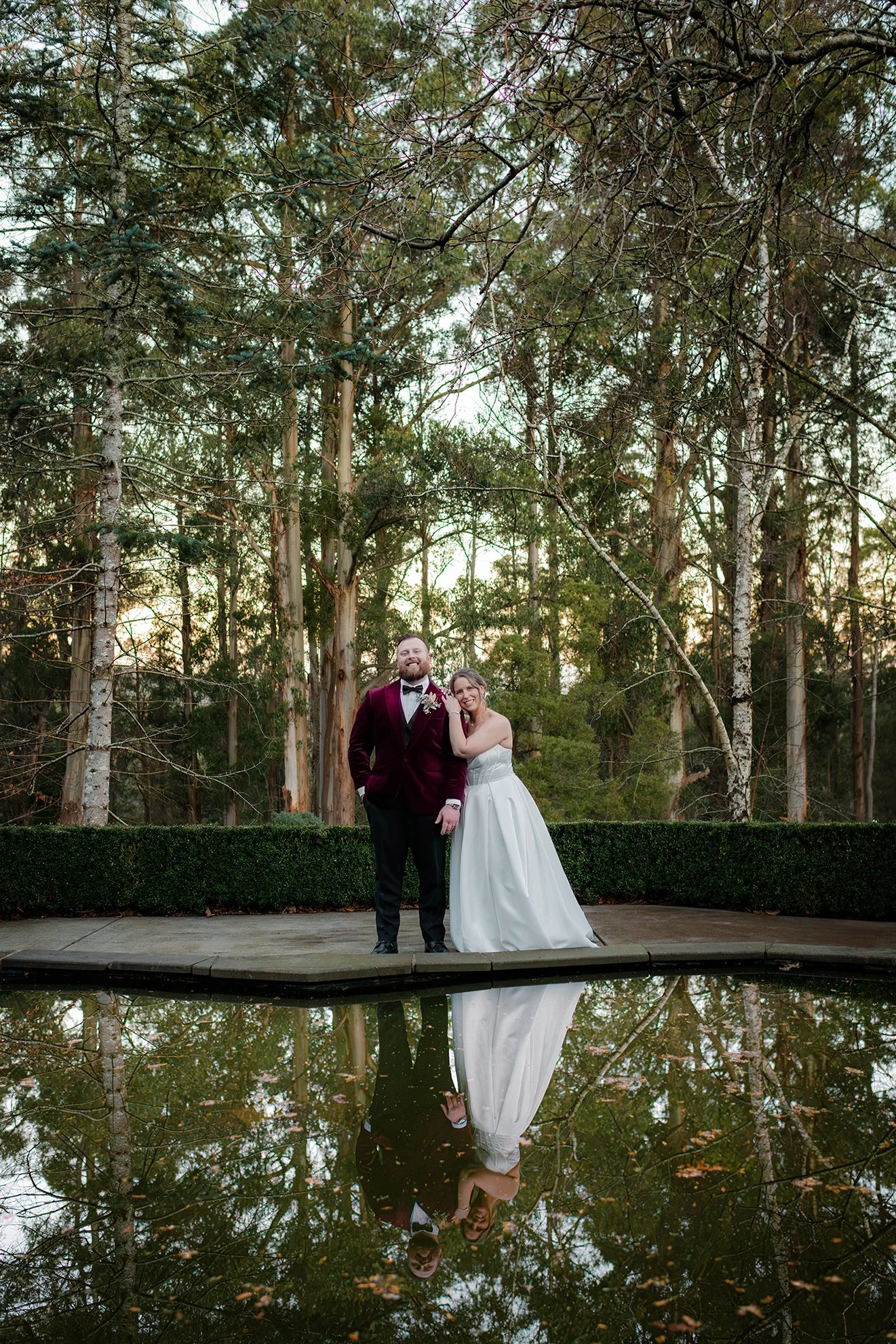 A bride and groom standing side by side outdoors in front of a forest, with their reflection visible in a pond.