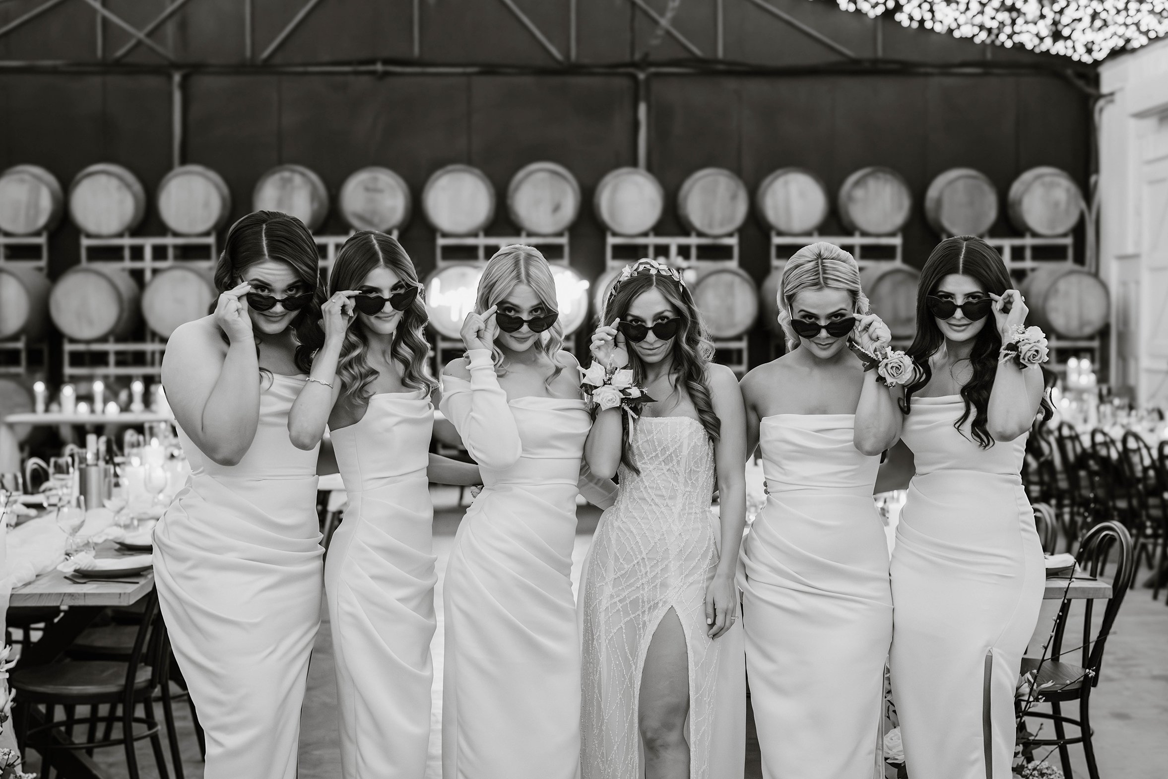 Seven women dressed in wedding or bridesmaid dresses, posing together while adjusting sunglasses, in a venue with wine barrels in the background, black and white photo.