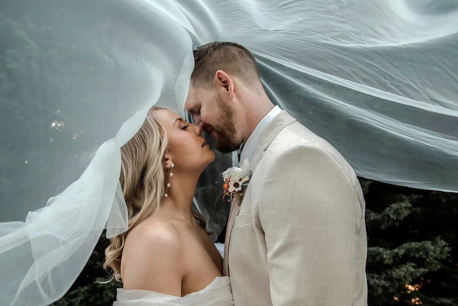 Bride and groom in wedding attire touching foreheads with a sheer veil flowing above them, outdoor setting with trees in the background.