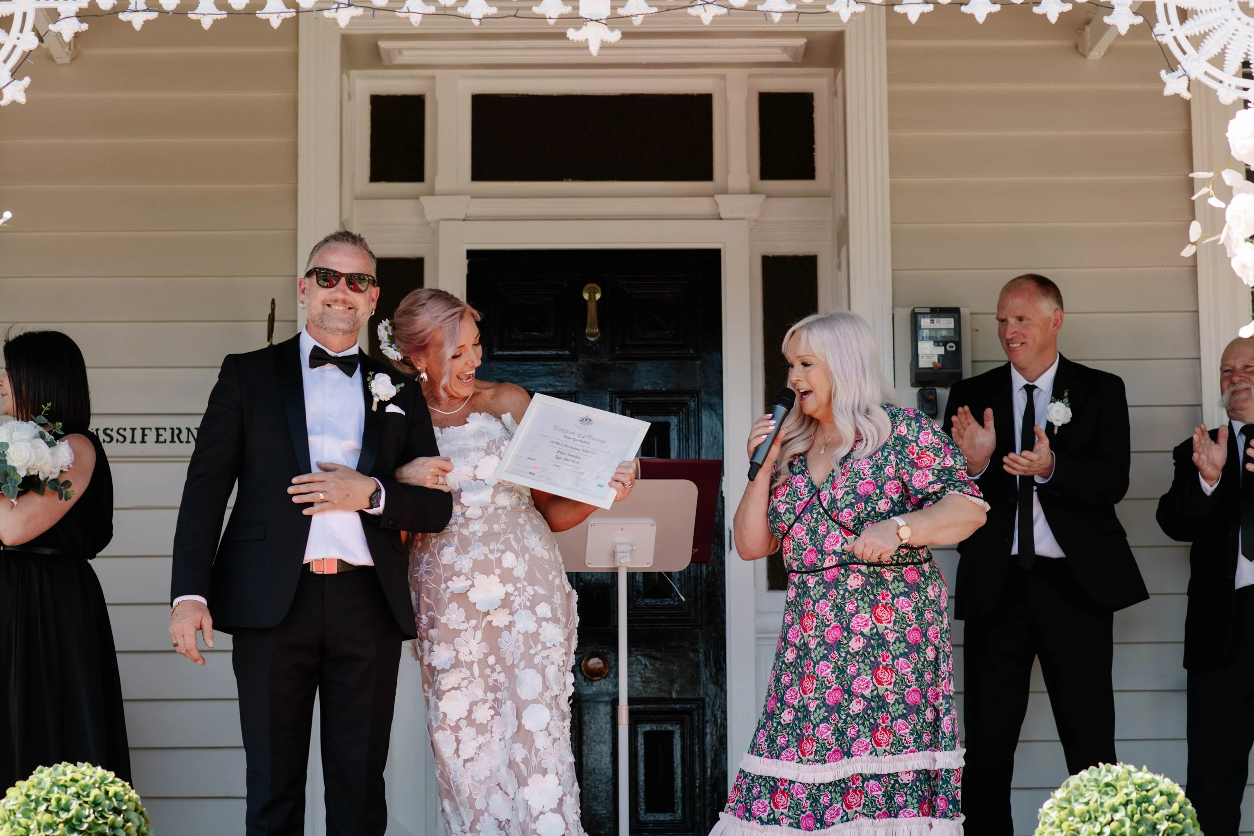 Bride holding up a marriage certificate beside the groom while the celebrant speaks and wedding party members applaud