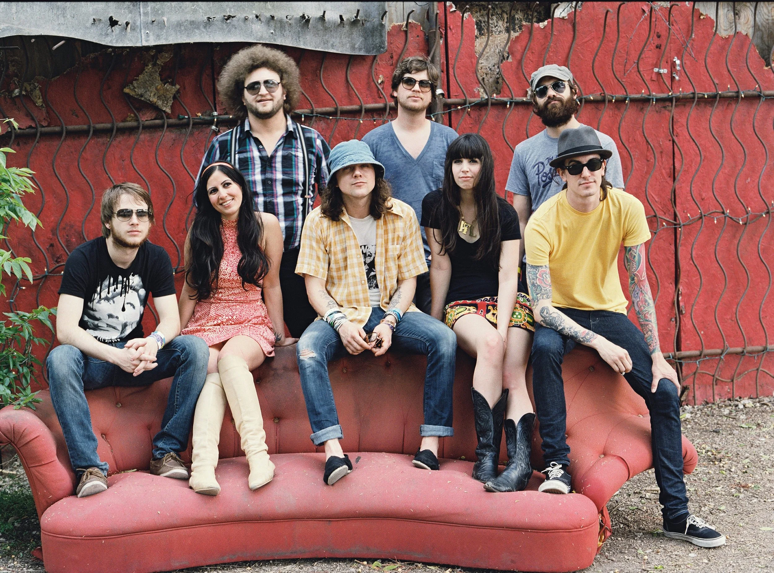 A group of nine young adults posing in front of a red wall with a rusted metal fence, sitting and standing on a vintage pink sofa outdoors, all wearing casual clothing and sunglasses.