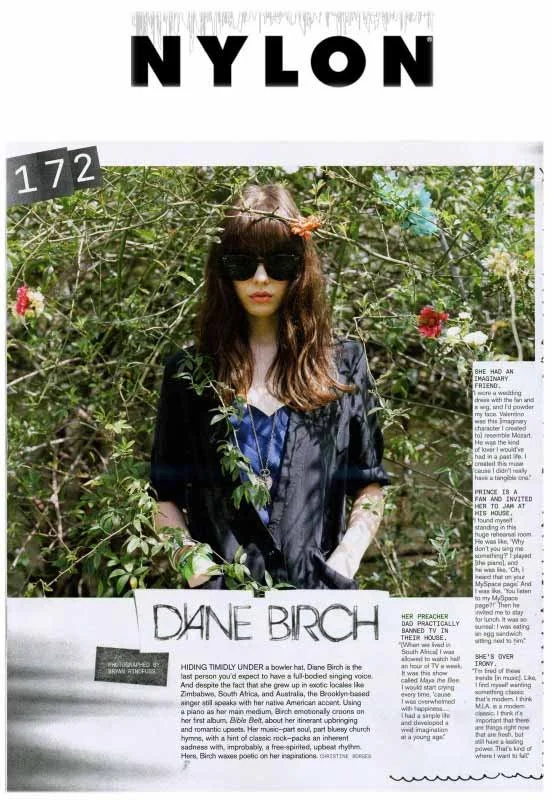 A woman with long brown hair, wearing sunglasses and a black cover-up, standing among green bushes and colorful flowers in a magazine cover photo.