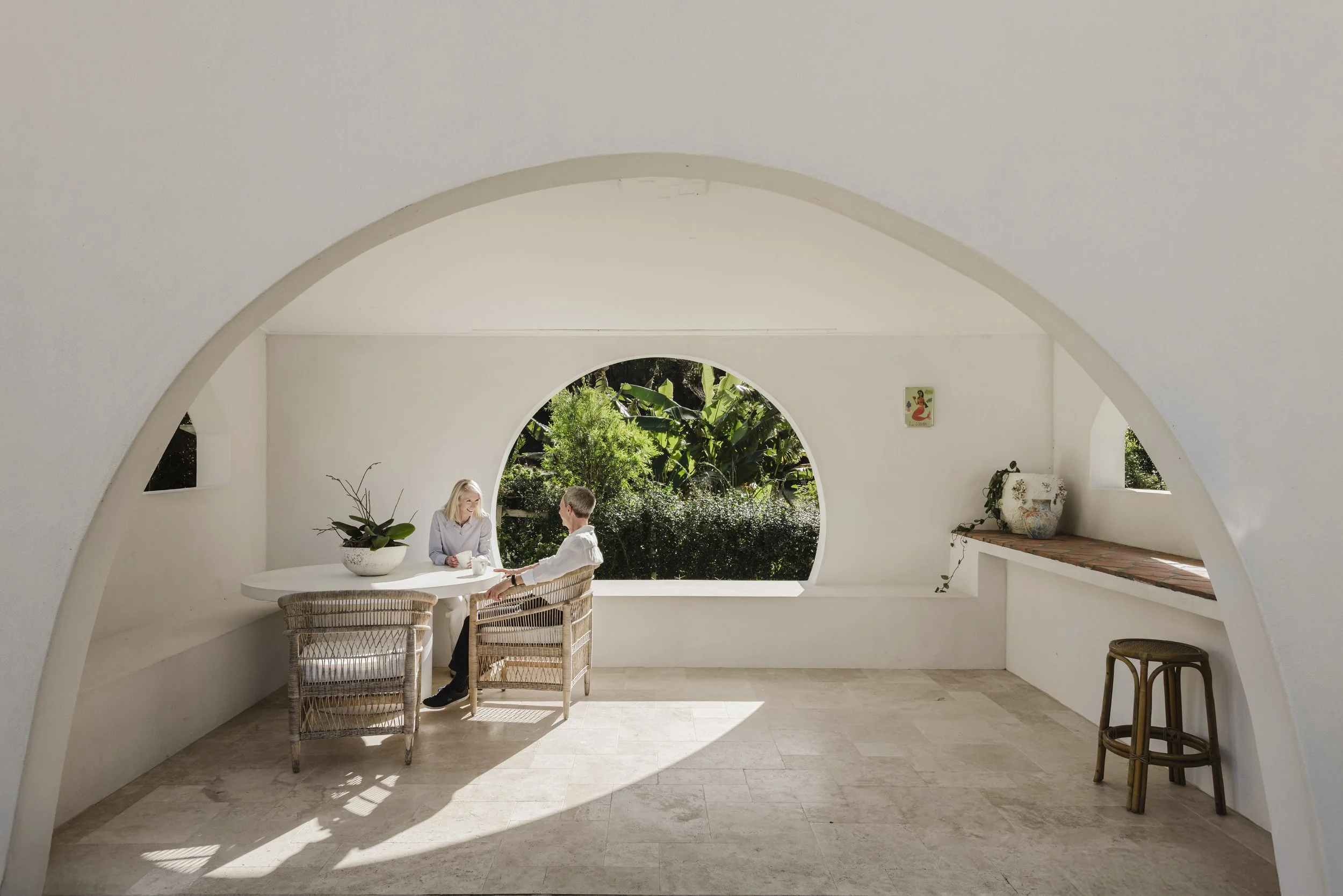 Two women sitting at a white table in a bright, minimalist room with rounded walls and a large round window showing greenery outside.