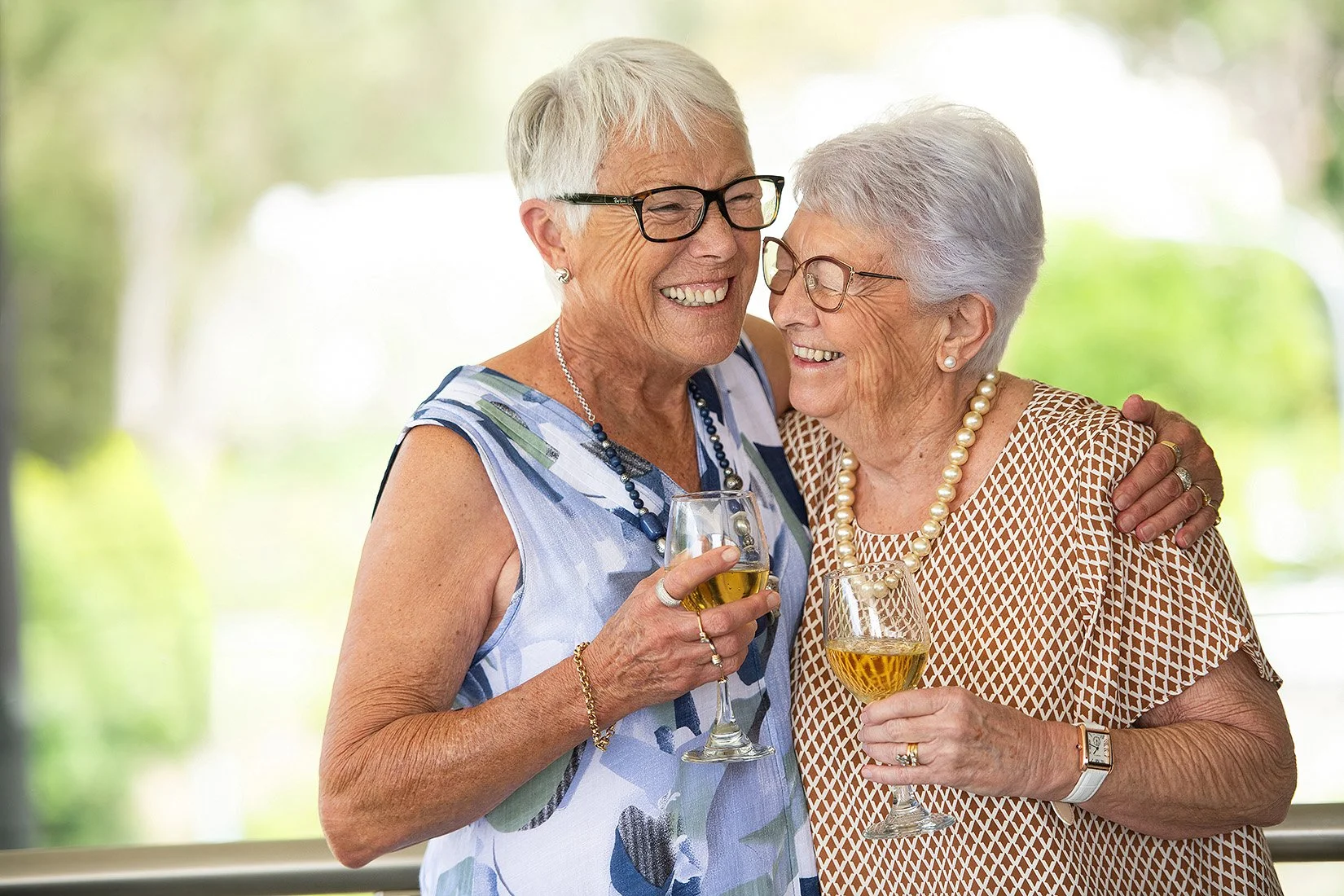 Two elderly women with gray hair and glasses smiling and hugging each other, holding glasses of white wine.