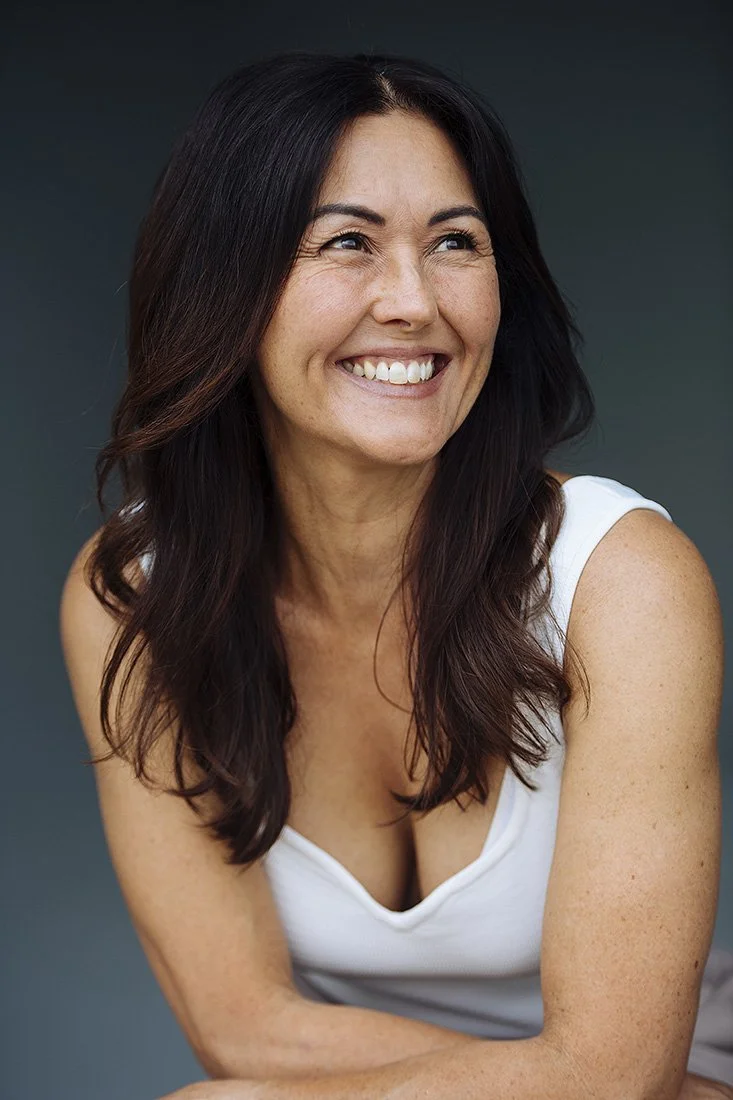 A woman with long dark hair smiling and looking to the side, wearing a white sleeveless top.