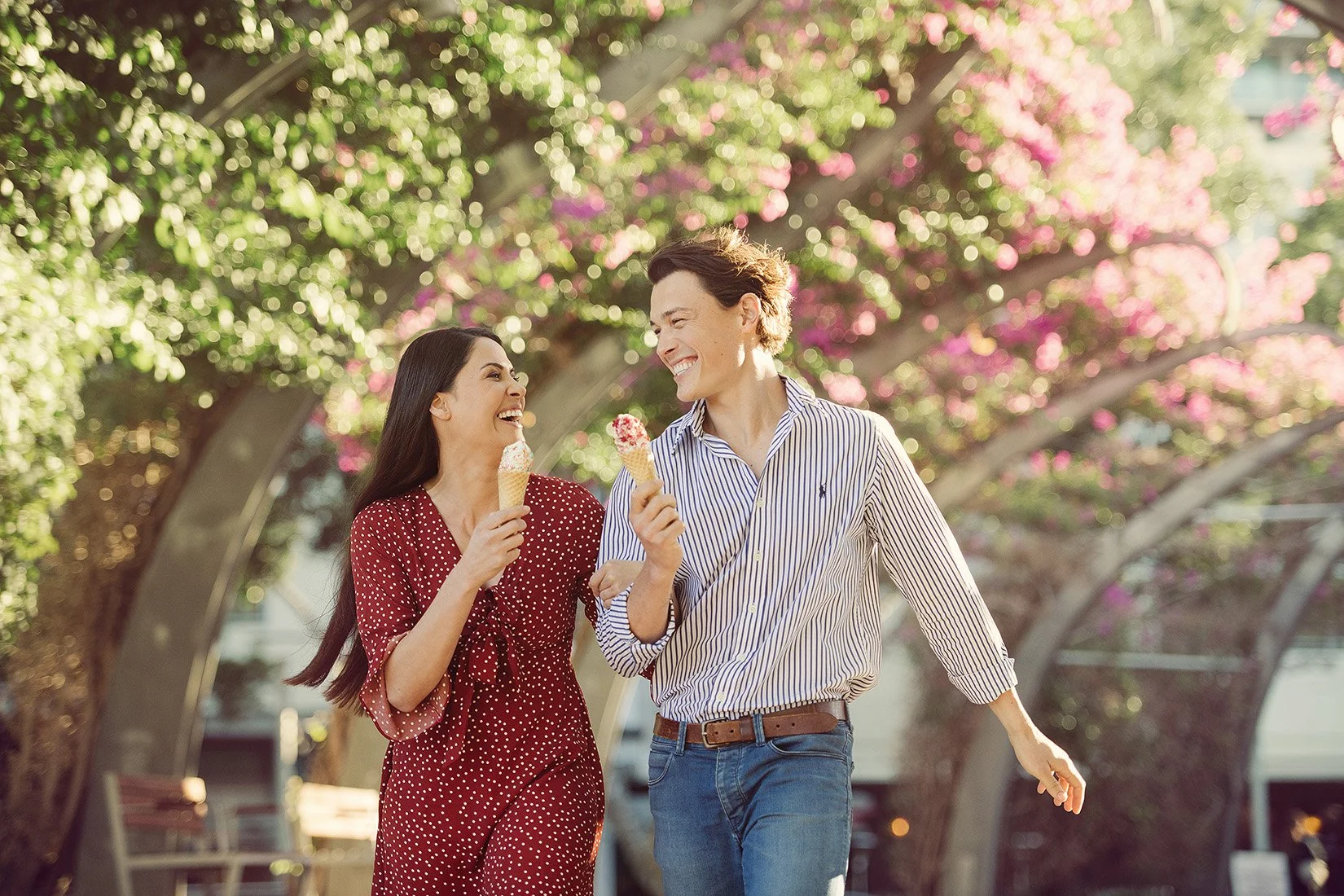 A joyful couple walking outdoors in a park, smiling and eating ice cream cones, with blooming pink and green trees in the background.