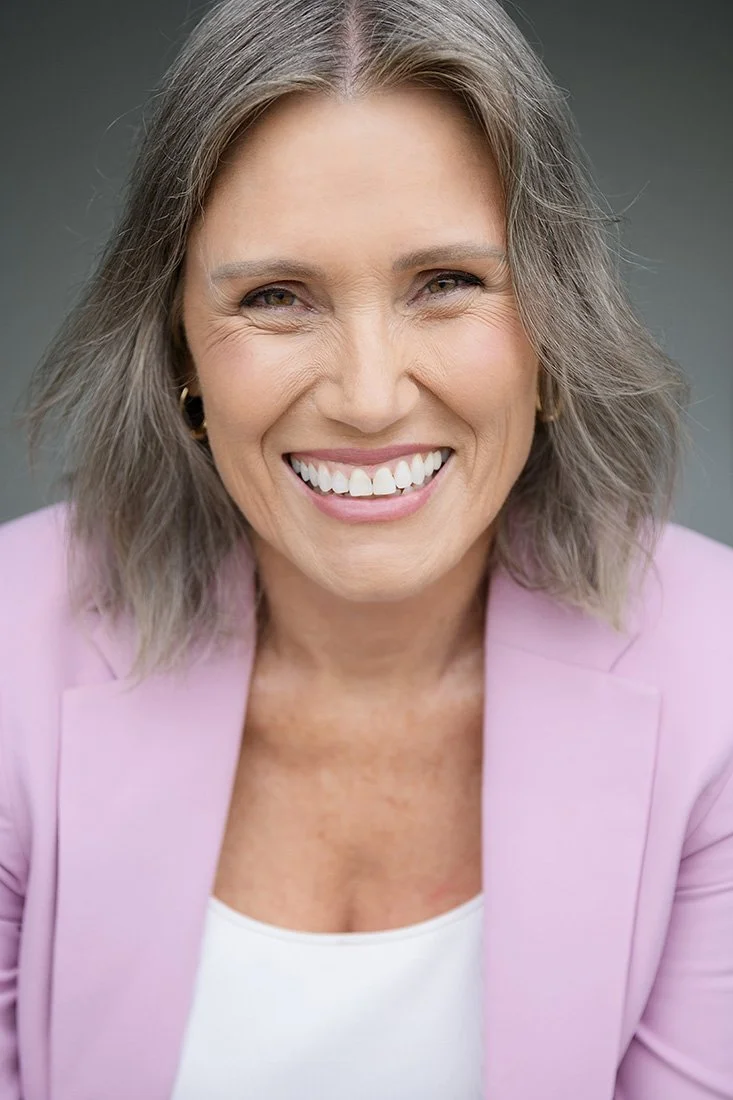 A smiling middle-aged woman with gray hair, wearing a pink blazer and white top, looking at the camera.