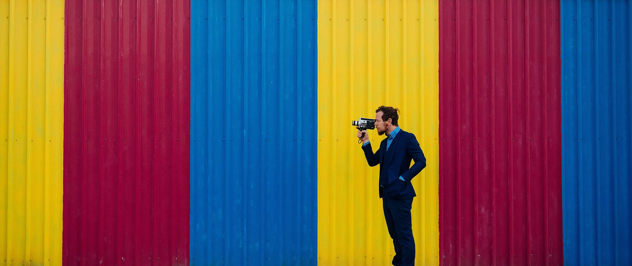 A man in a navy blue suit taking a photograph with a vintage camera against a colorful corrugated metal wall with vertical red, yellow, and blue stripes.