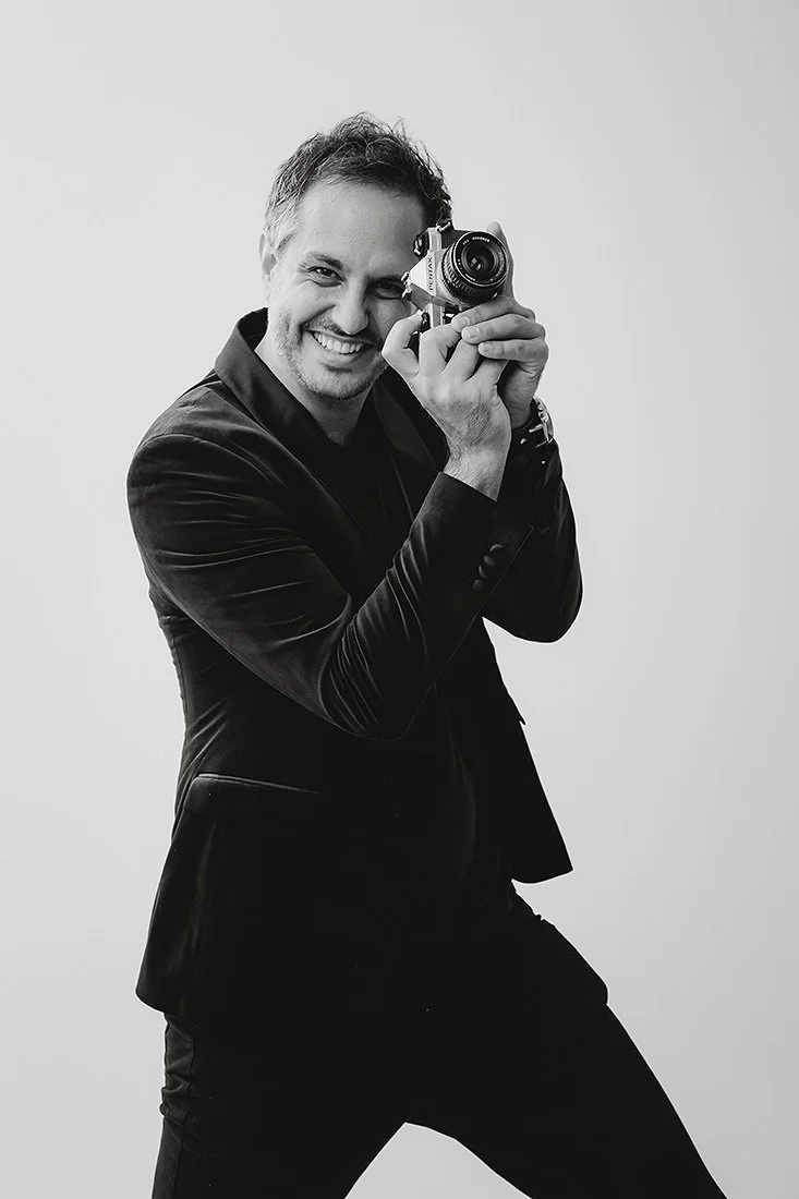 Jack Venables cheekily smiling and holding a camera up to his face, wearing a dark suit, posing against a plain background.