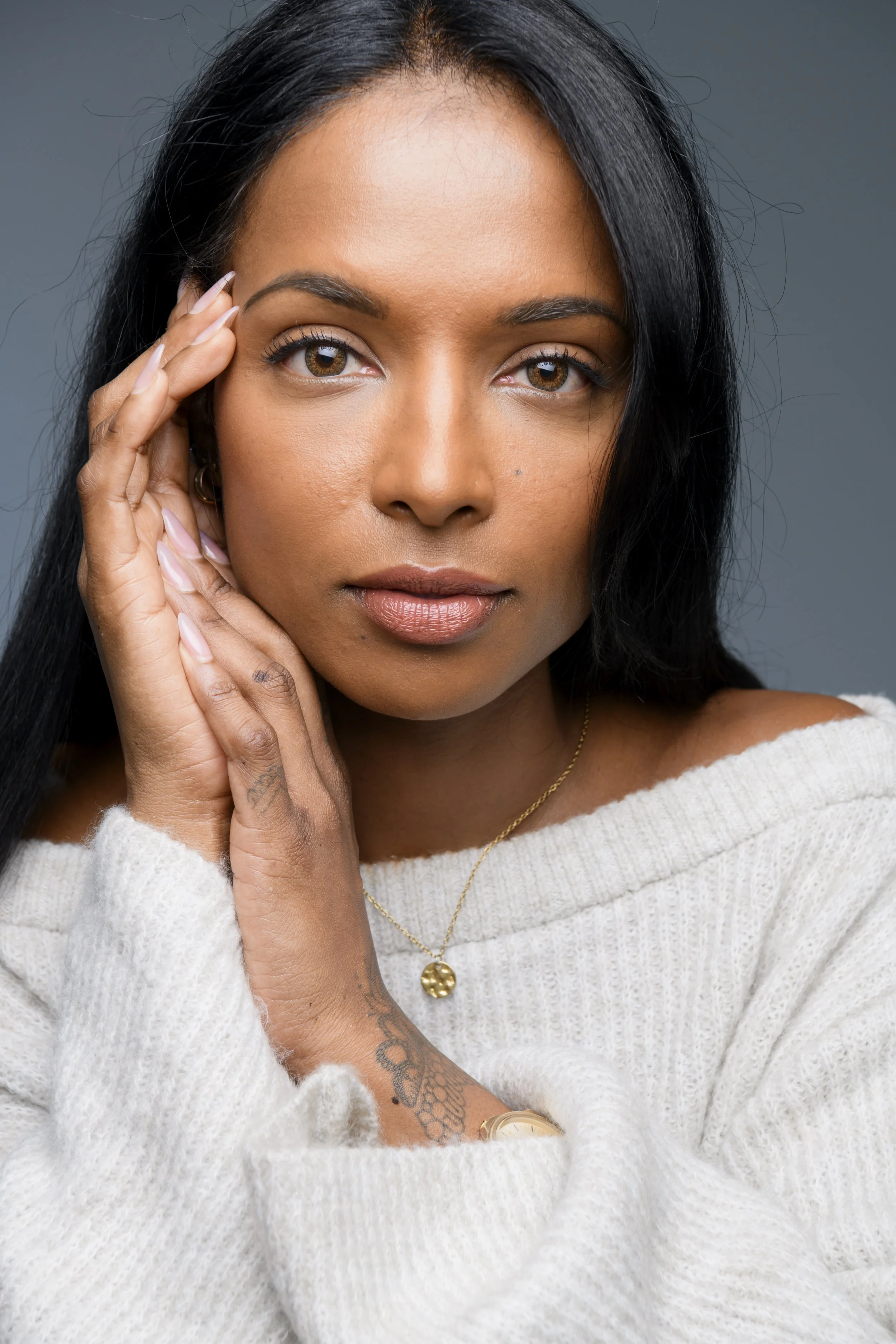 Close-up portrait of a woman with black hair, brown eyes, wearing a white sweater, gold necklace, and watch, touching her face with one hand, looking directly at the camera.