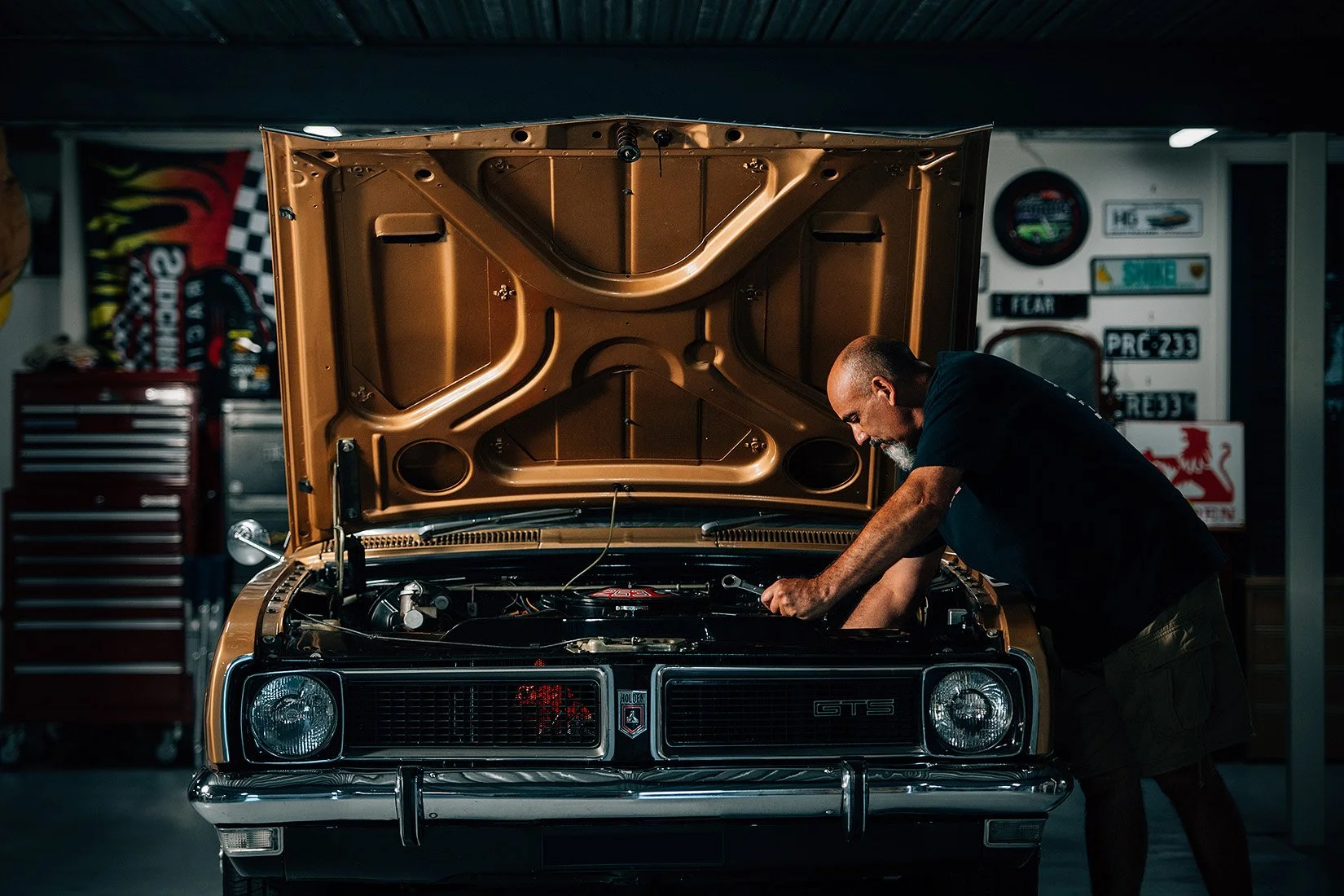 A man working on the engine of a vintage gold-colored Pontiac GTS car in a garage, with the hood open and racing or car enthusiast signs on the wall behind him.