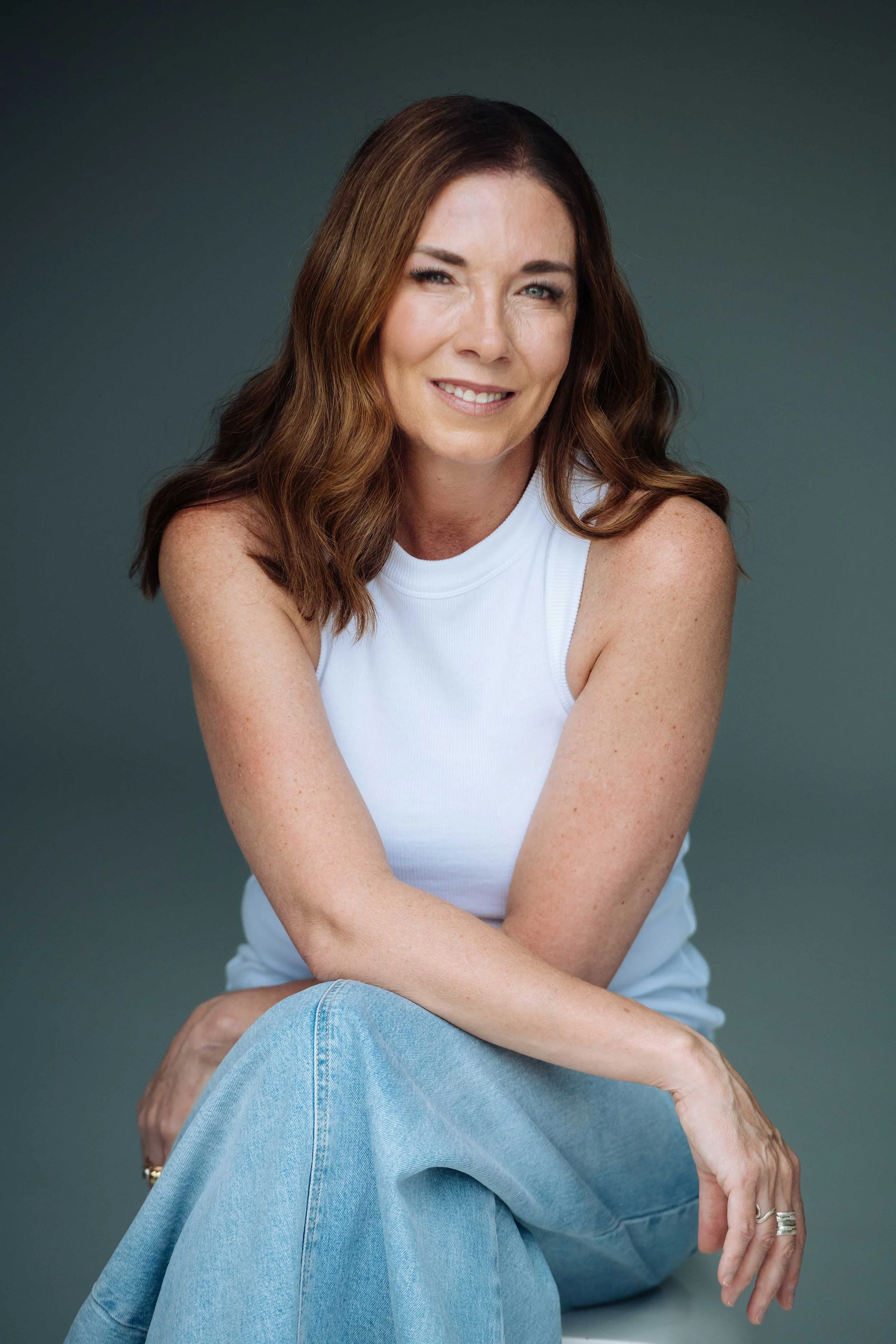 A woman with wavy brown hair and fair skin sitting against a plain background, wearing a white sleeveless top and light blue jeans, smiling and winking at the camera.