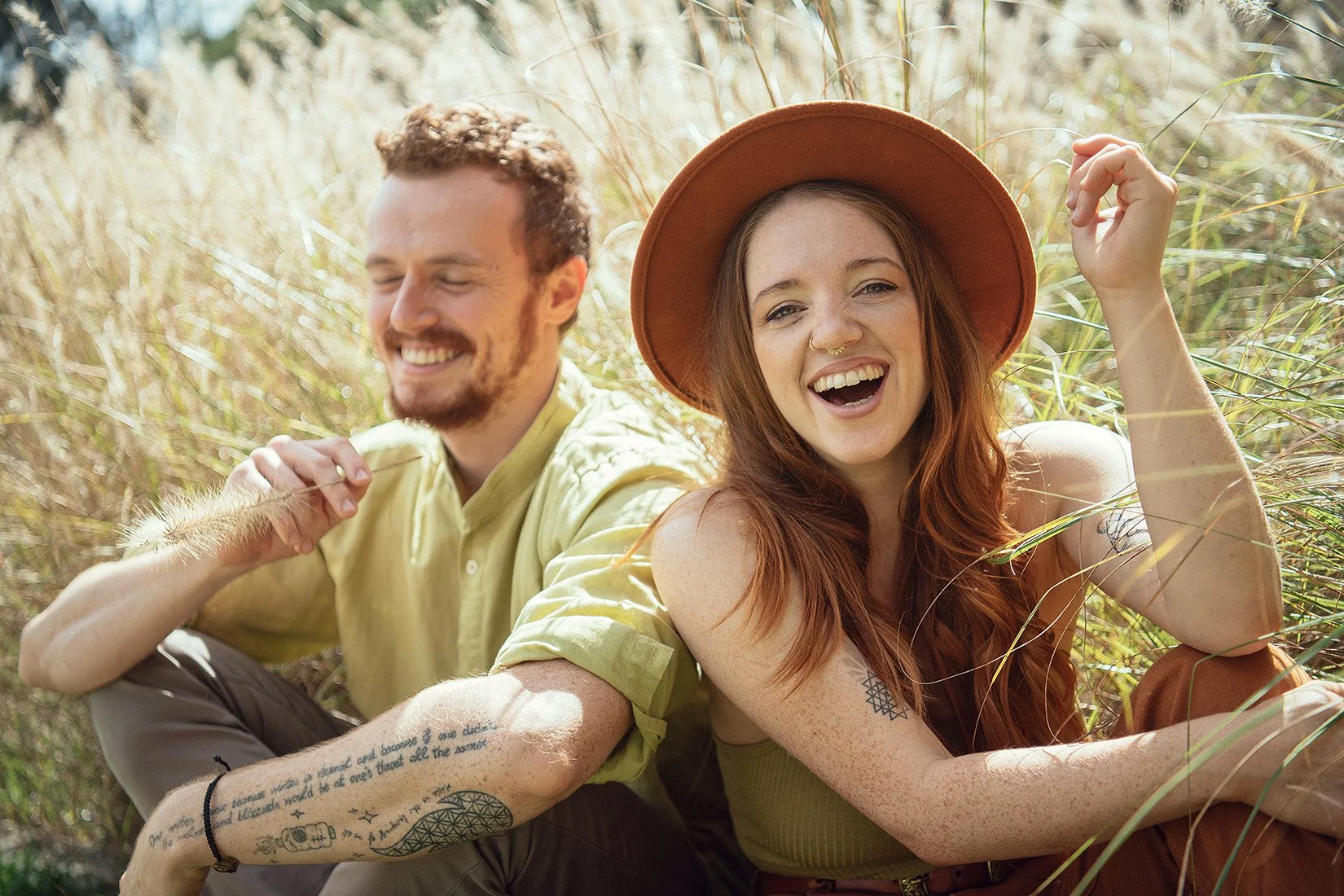 A smiling young woman with long red hair, a wide-brimmed hat, and tattoos on her arm, sitting outdoors in a grassy field, beside a young man with curly hair and a beard, both enjoying a sunny day.