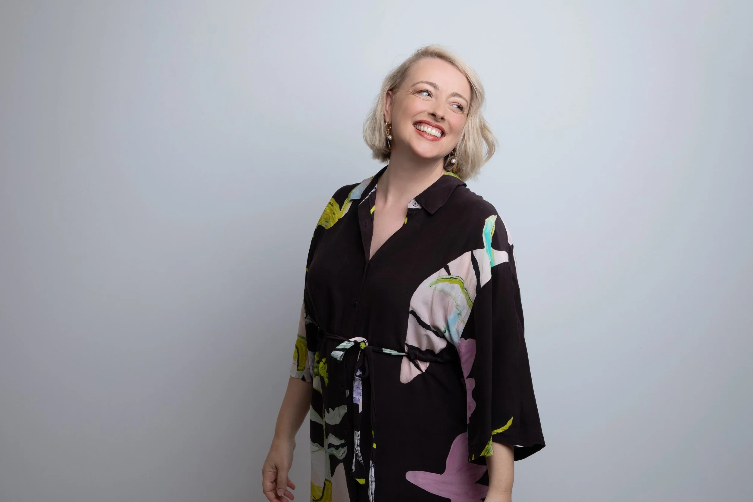 Helen Stump with blonde hair, wearing a black dress with colourful floral patterns, looks to her right in front of a plain white background.
