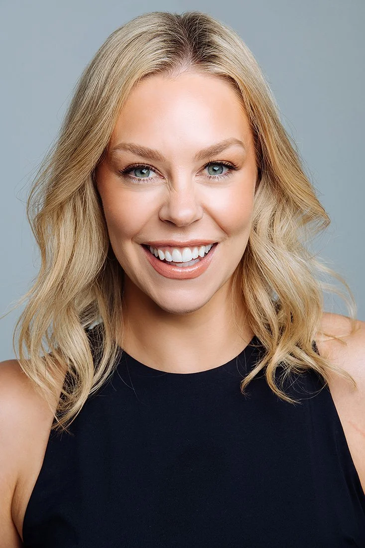 A young woman with blonde wavy hair, blue eyes, and fair skin smiling while wearing a black sleeveless top.