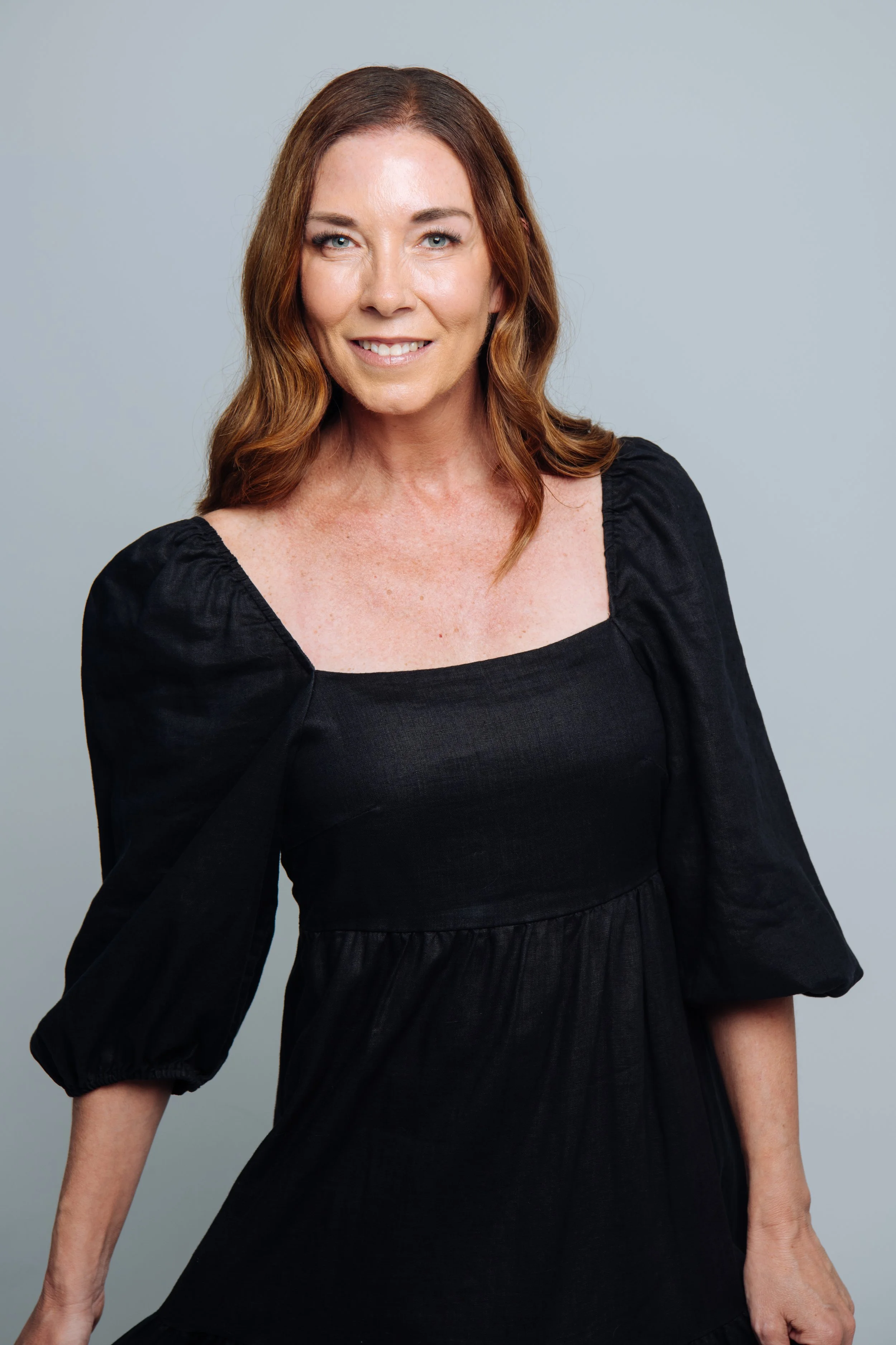 A woman with red hair and blue eyes wearing a black dress with puffed sleeves, standing against a light gray background.