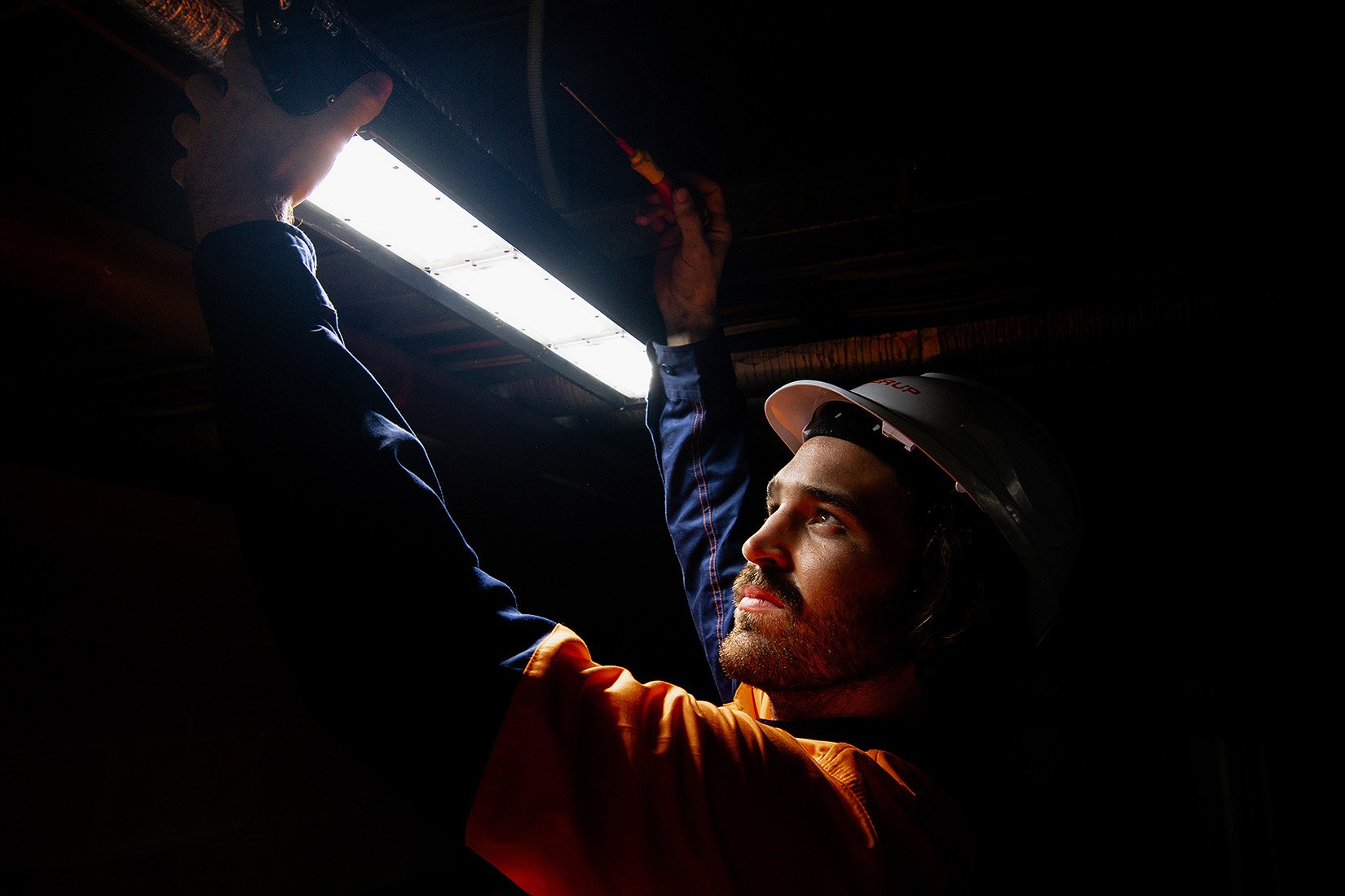 Worker installing or inspecting a light fixture on a dark ceiling, wearing a helmet and safety gear.