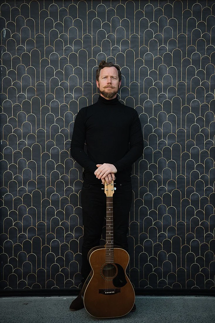 A man with a beard and mustache wearing a black turtleneck and black pants standing in front of a patterned dark wall, holding an acoustic guitar vertically in front of him.
