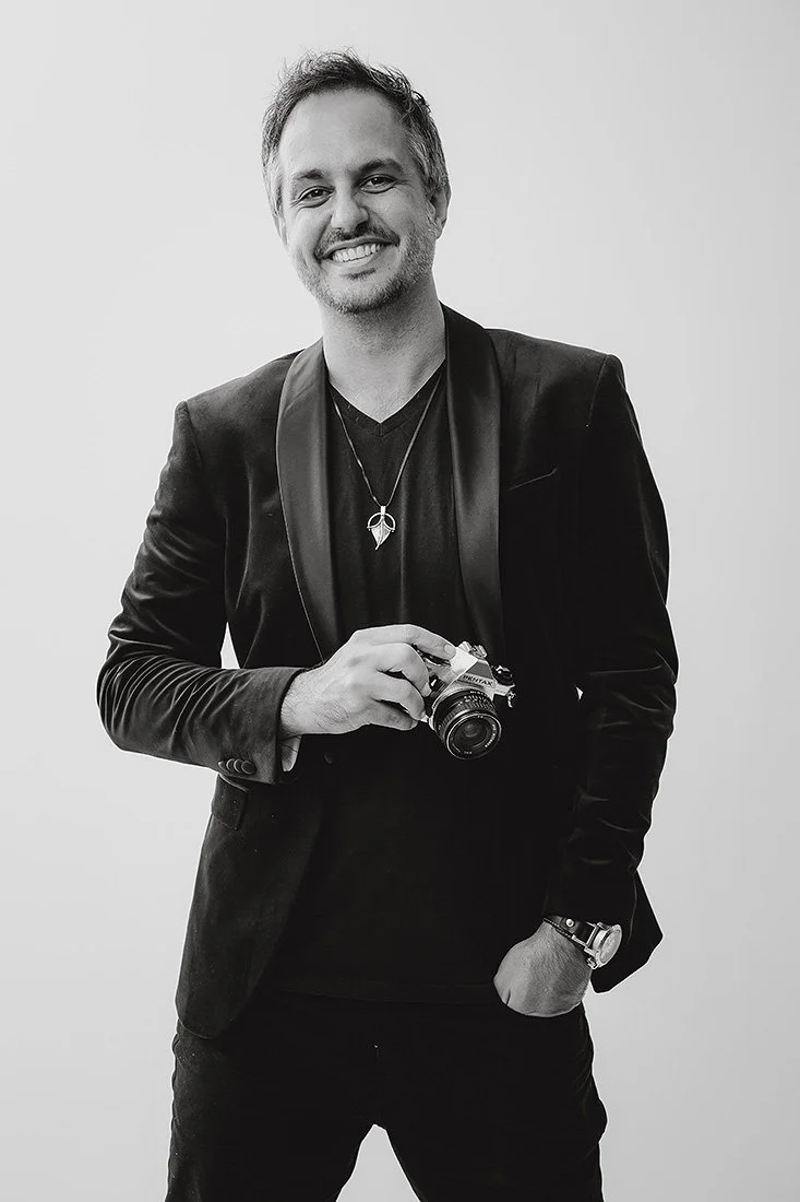 Black and white photo of a smiling man with short hair, wearing a black blazer with a satin lapel, a dark shirt, and a wristwatch, holding a camera, standing against a plain background.