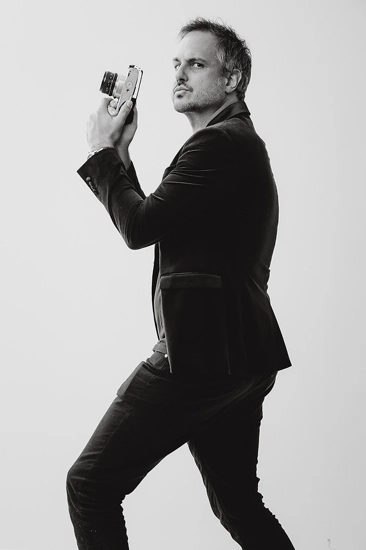 Jack Venables a man who looks remarkably like Mark Ruffalo with short hair and a beard holding a camera, standing in silly pose against a plain background.