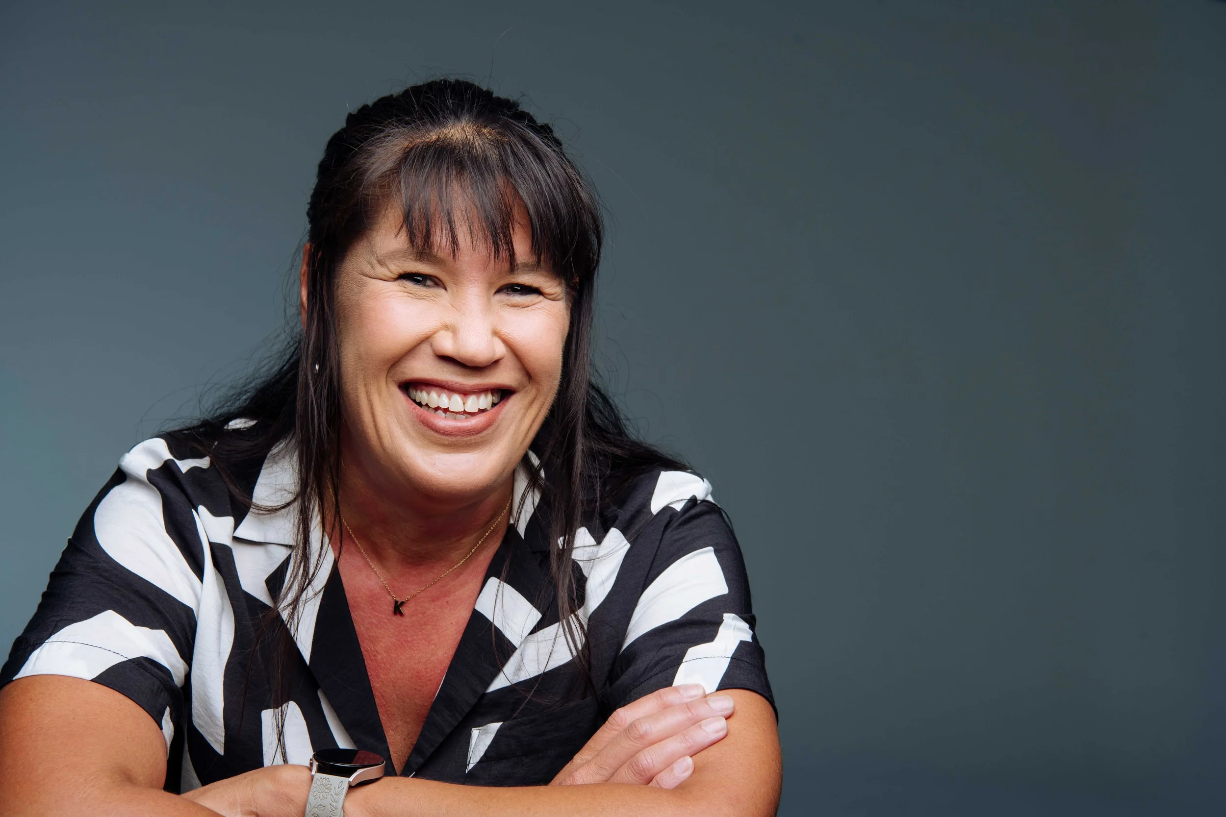Smiling woman with dark hair and bangs, wearing a black and white patterned shirt, sitting with arms crossed against a dark grey background.