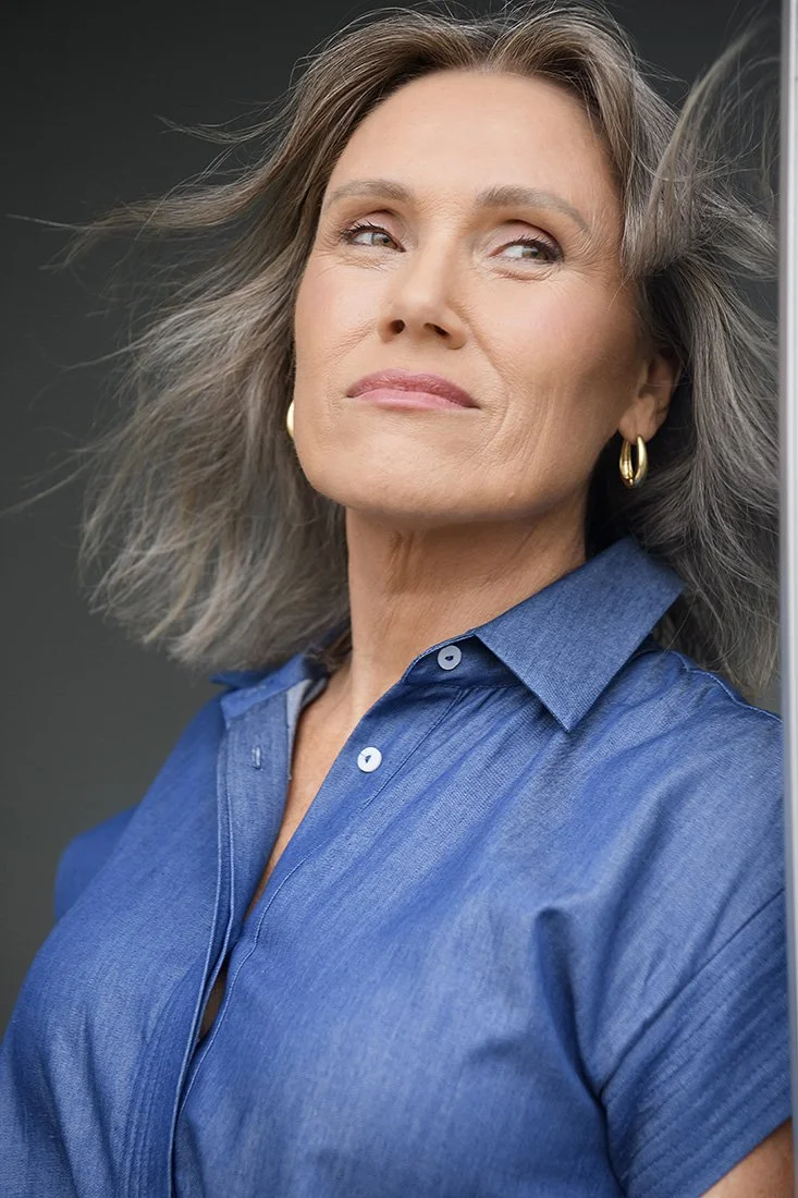 A mature woman with gray hair, wearing a blue button-up shirt and gold hoop earrings, stands with a contemplative expression against a dark background.