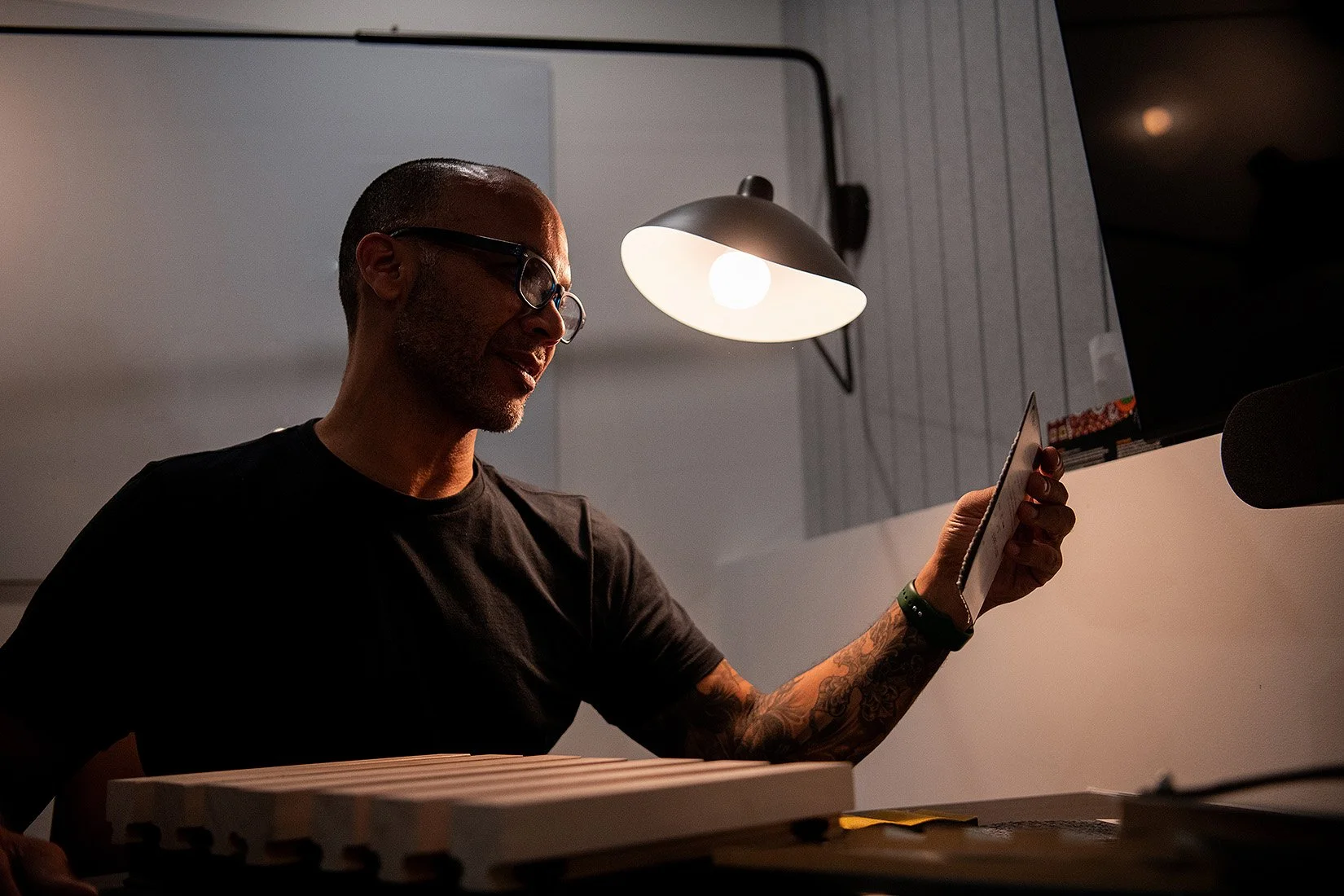 A man with glasses, a tattooed arm, and a black t-shirt sitting at a desk, looking at his phone in a dimly lit room illuminated by a desk lamp and a computer monitor.