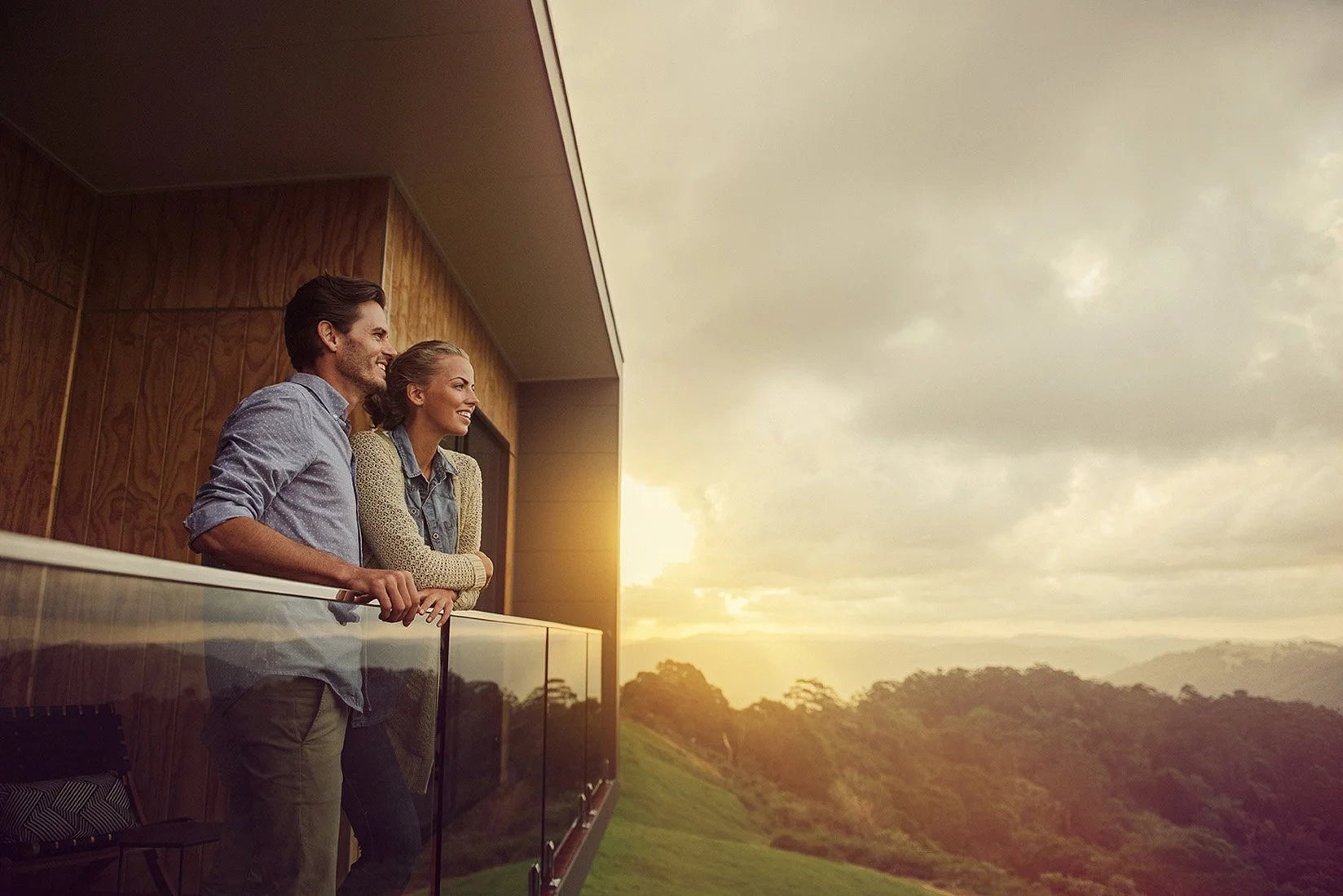 A man and woman standing on a balcony, looking at the sunset over a landscape of hills and trees.