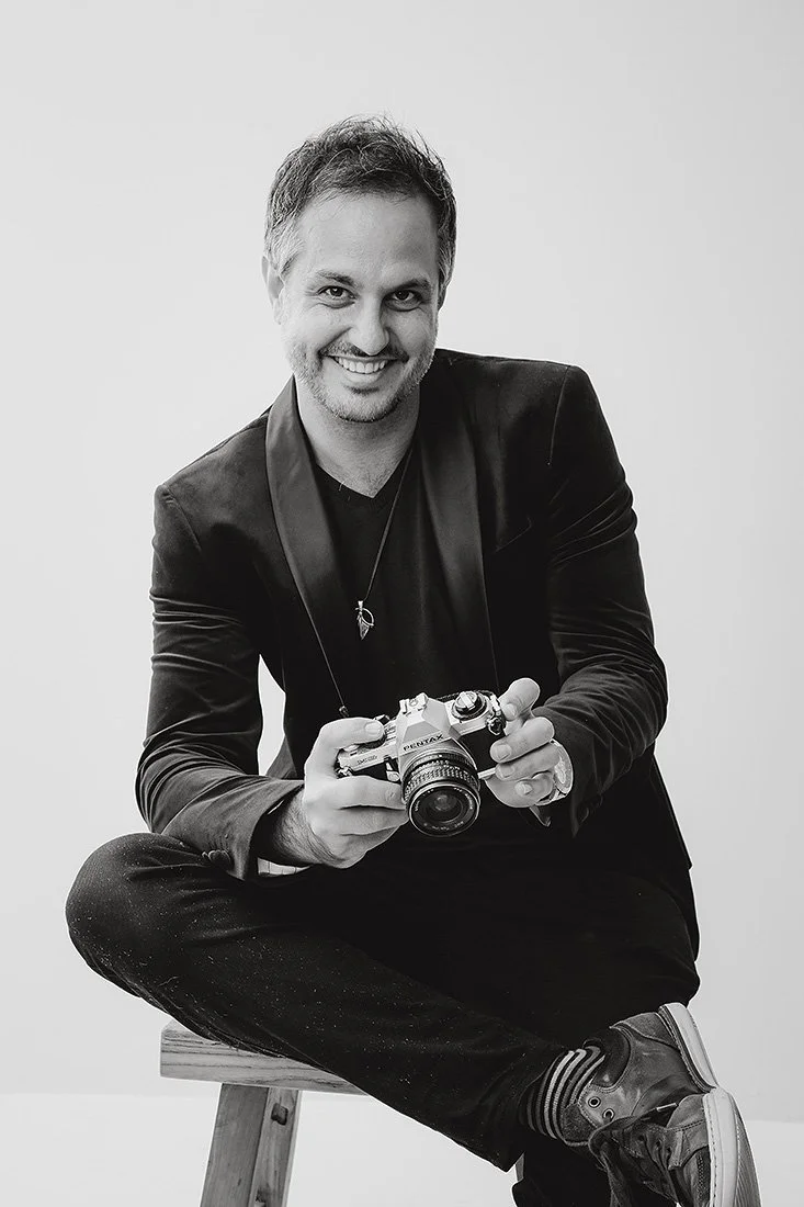 Jack Venables, sitting cross-legged on a stool, holding a vintage camera, dressed in a dark blazer, black pants, and sneakers, in a studio setting with a plain background.
