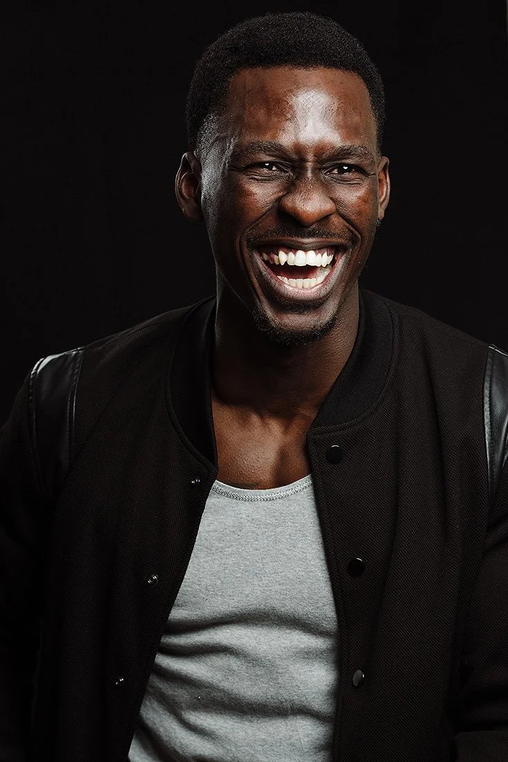 A smiling man with dark skin, short black hair, wearing a black jacket over a light gray shirt, against a black background.