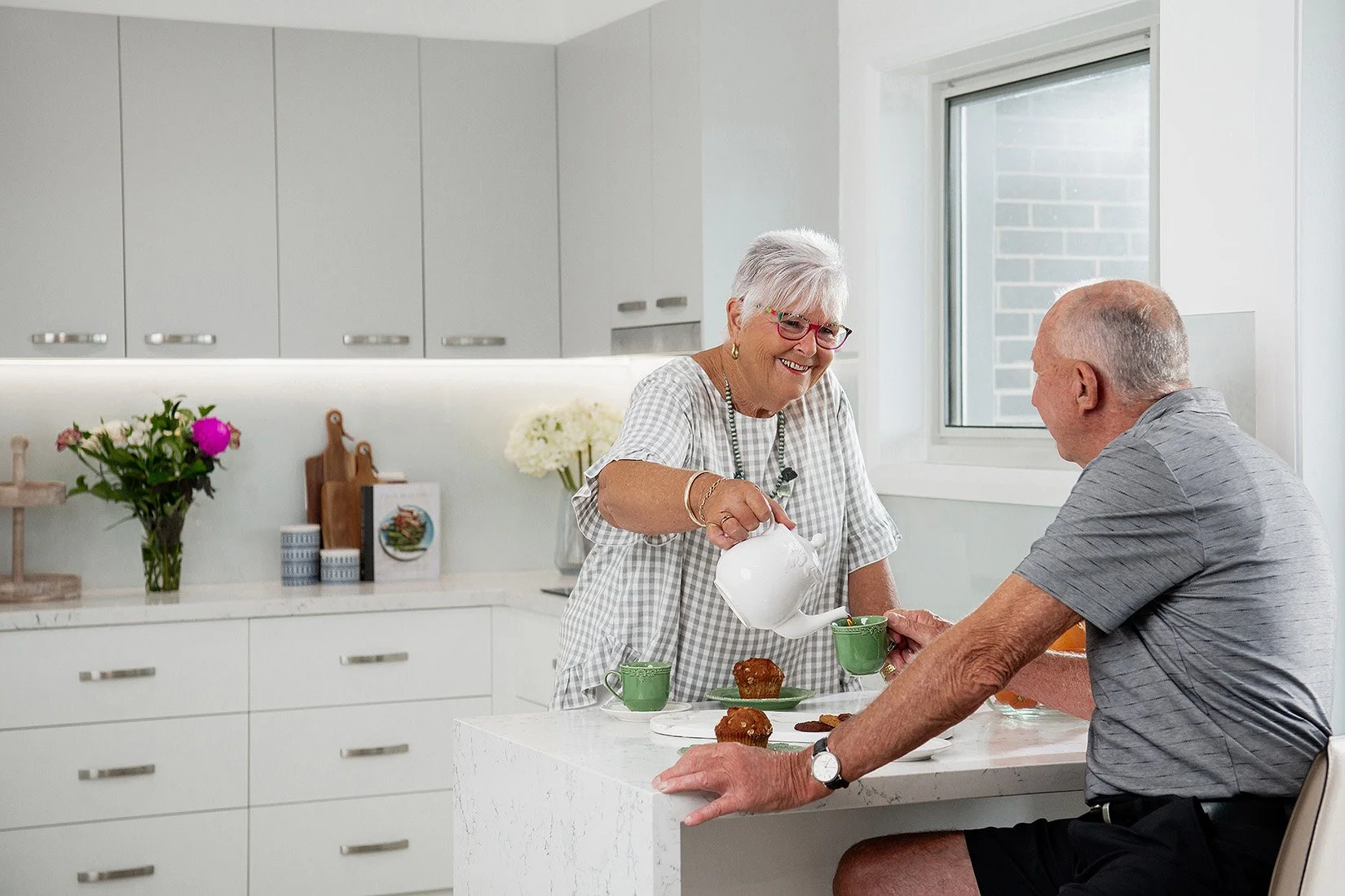 An elderly woman pouring tea into a green cup for an elderly man at a kitchen island.